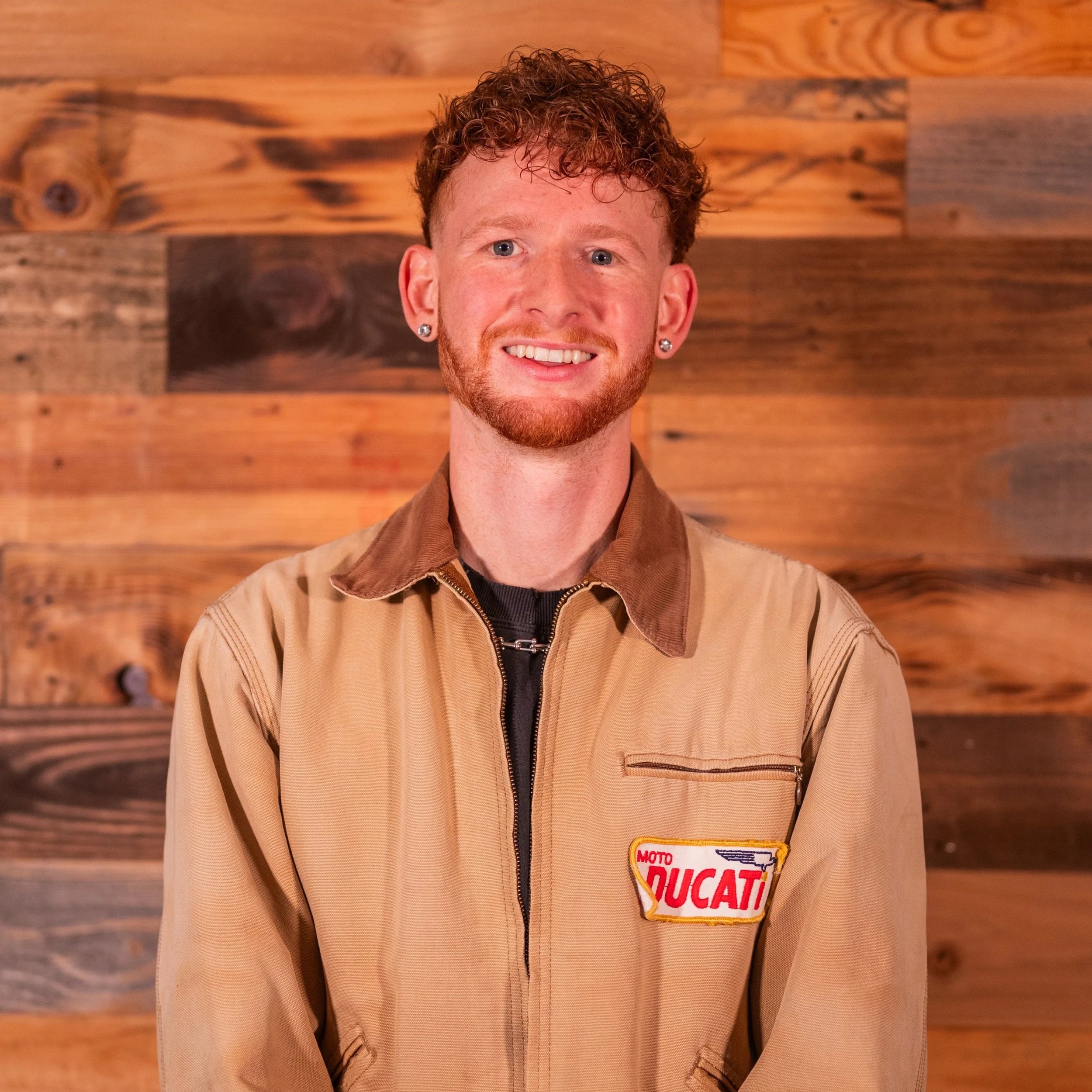 Smiling person with curly hair, wearing earrings and a white shirt, standing against a geometric background.