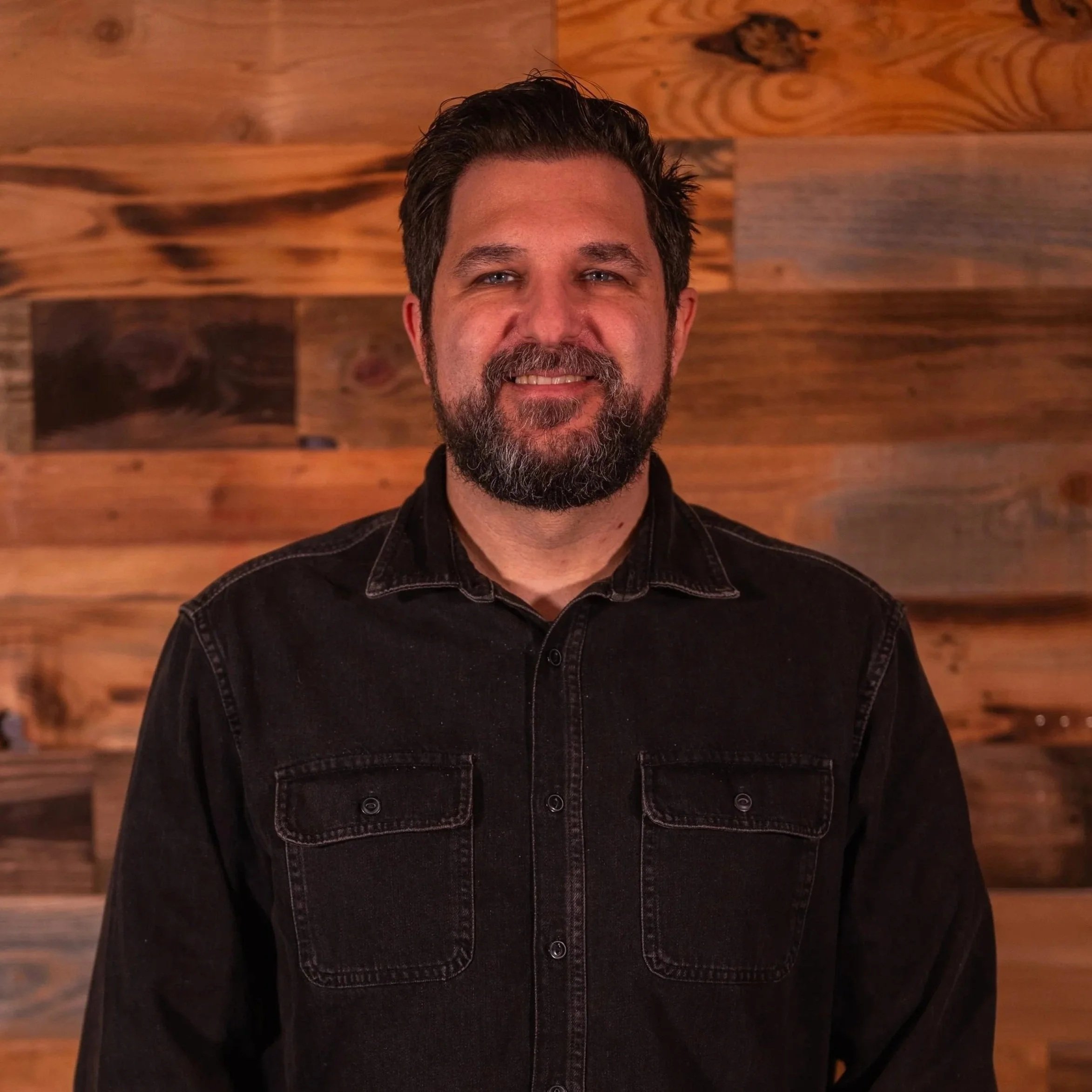 Man with a beard and plaid shirt smiling in front of a tree trunk.