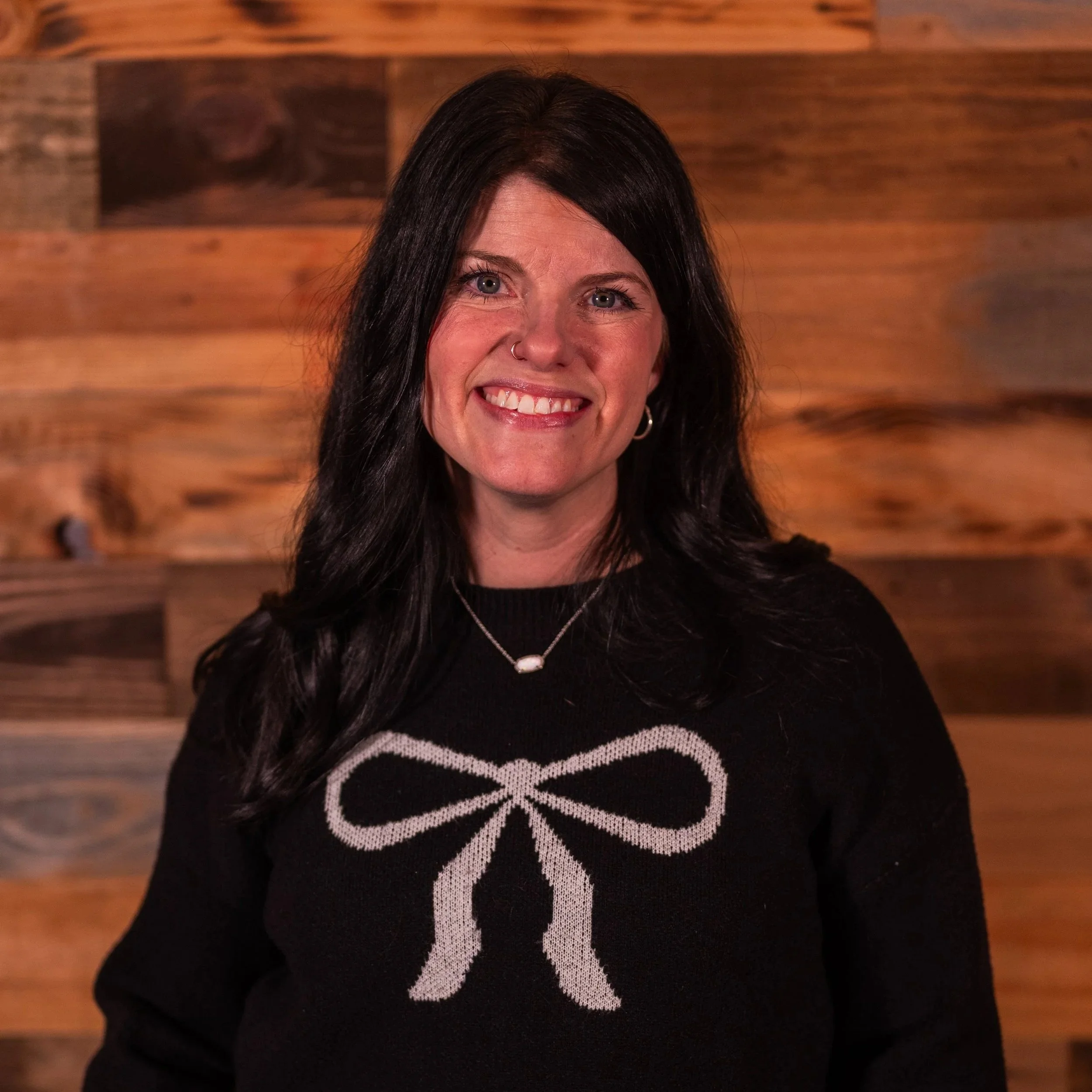 Smiling woman with dark hair standing in front of a large tree, wearing a brown sweater and black top, outdoors.