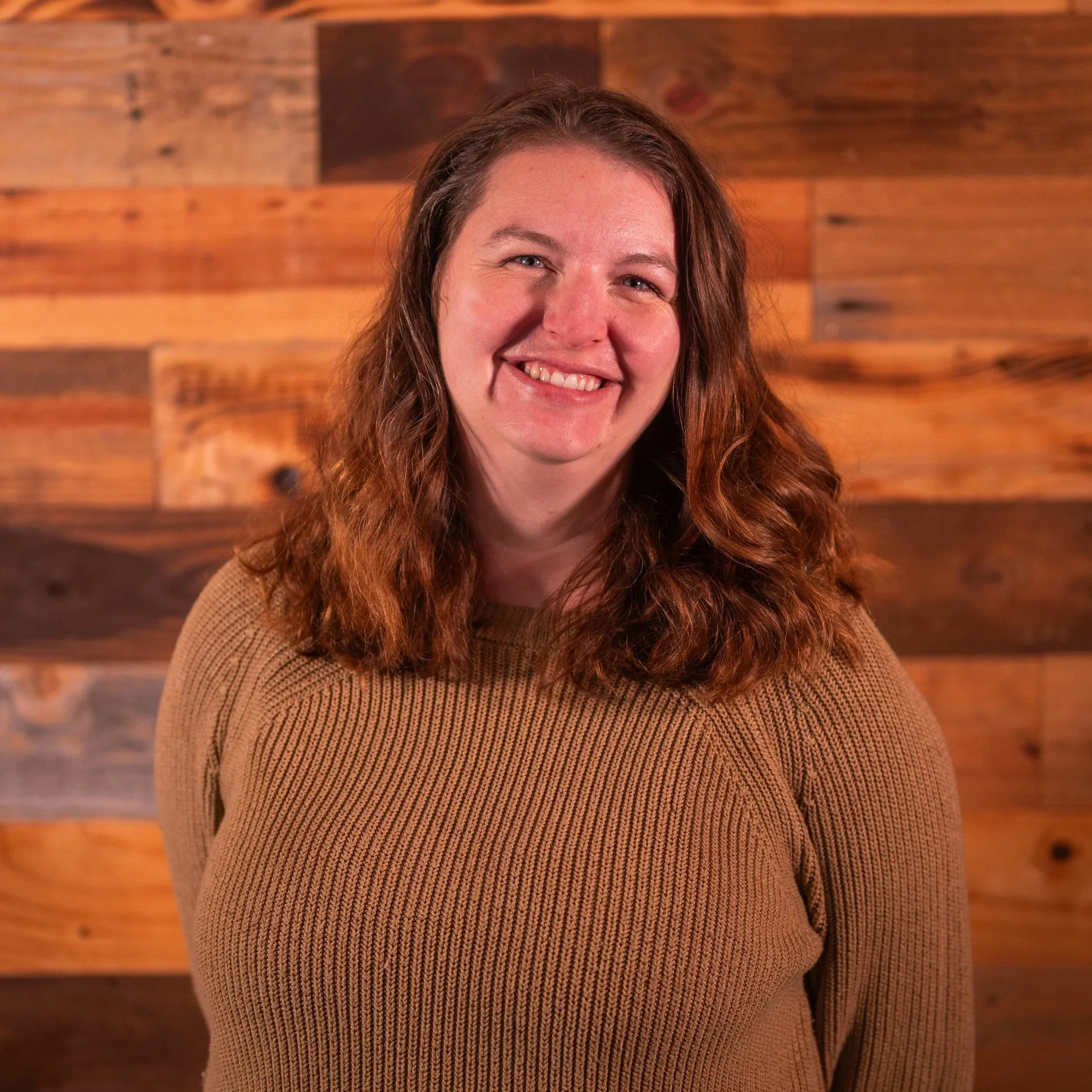 Smiling person with long hair, wearing a dark top, standing in front of a geometric-patterned background.