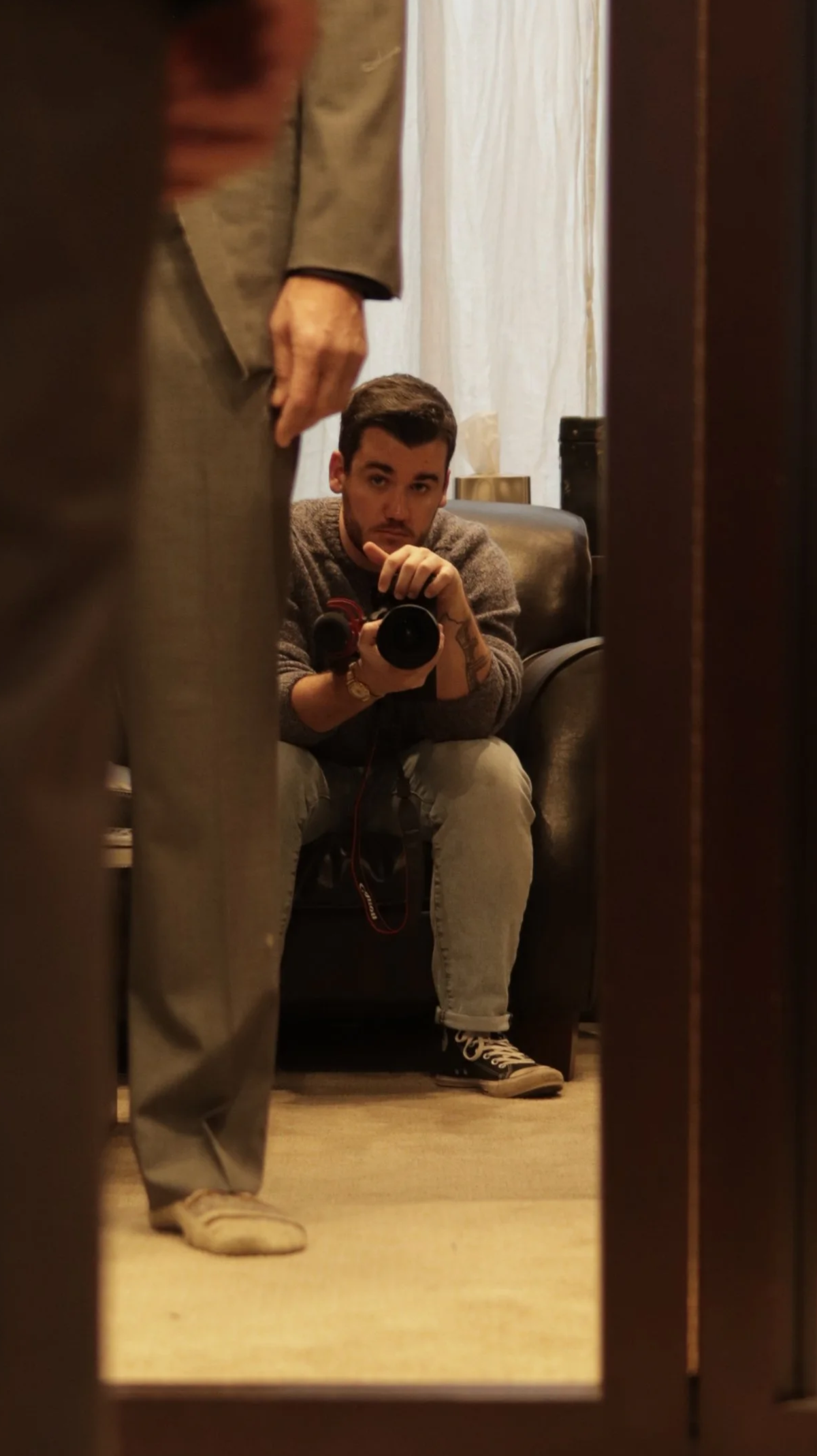 A man smiling, sitting on a chair, in a filming or photography studio with camera equipment and a dark background, illuminated by warm lighting and decorative lamps.