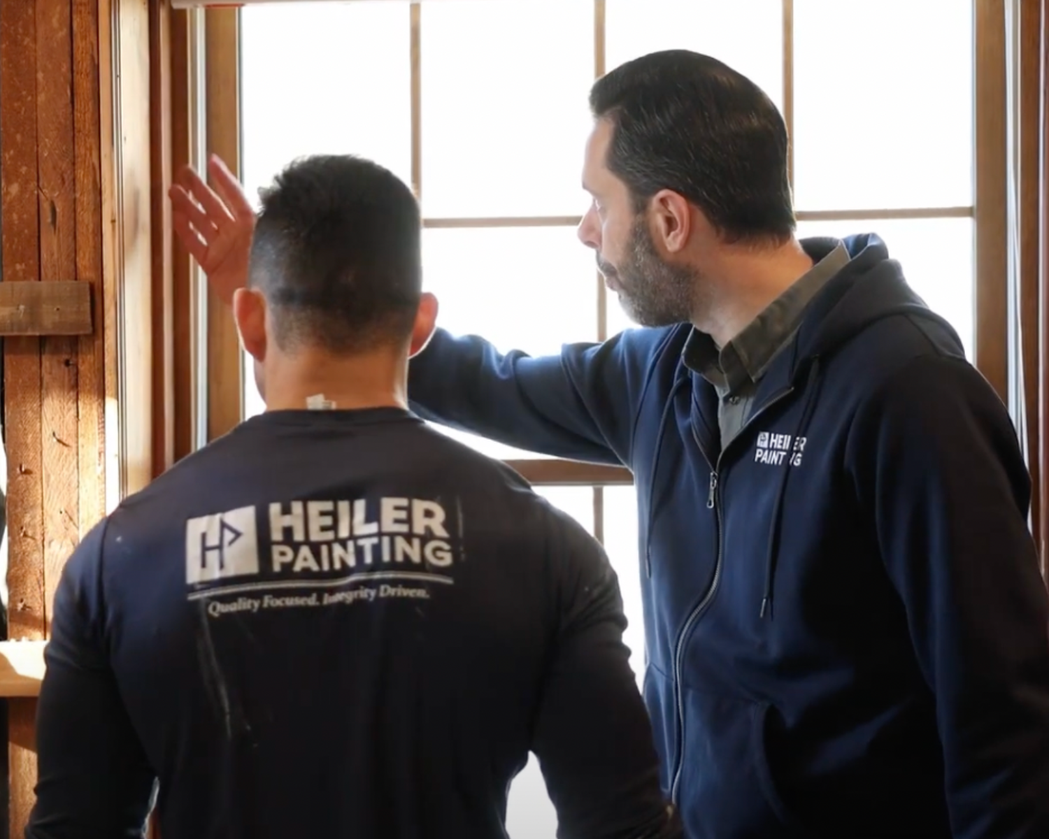 Two men in dark blue hoodies with 'HEILER PAINTING' logo are inside a wooden building, one looking at a window and the other gesturing towards it, possibly discussing a painting or renovation project.