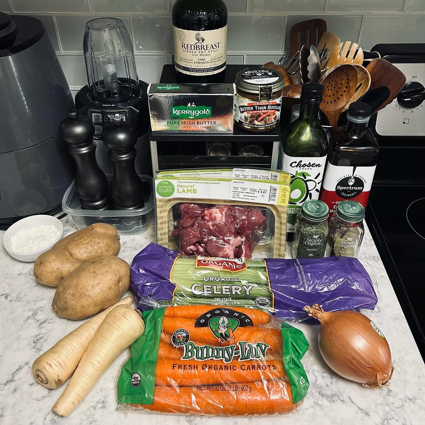 Assorted ingredients on a kitchen counter, including potatoes, parsnips, carrots, an onion, beef stew meat, celery, shredded cheese, herbs, a bottle of whiskey, and various cooking utensils.