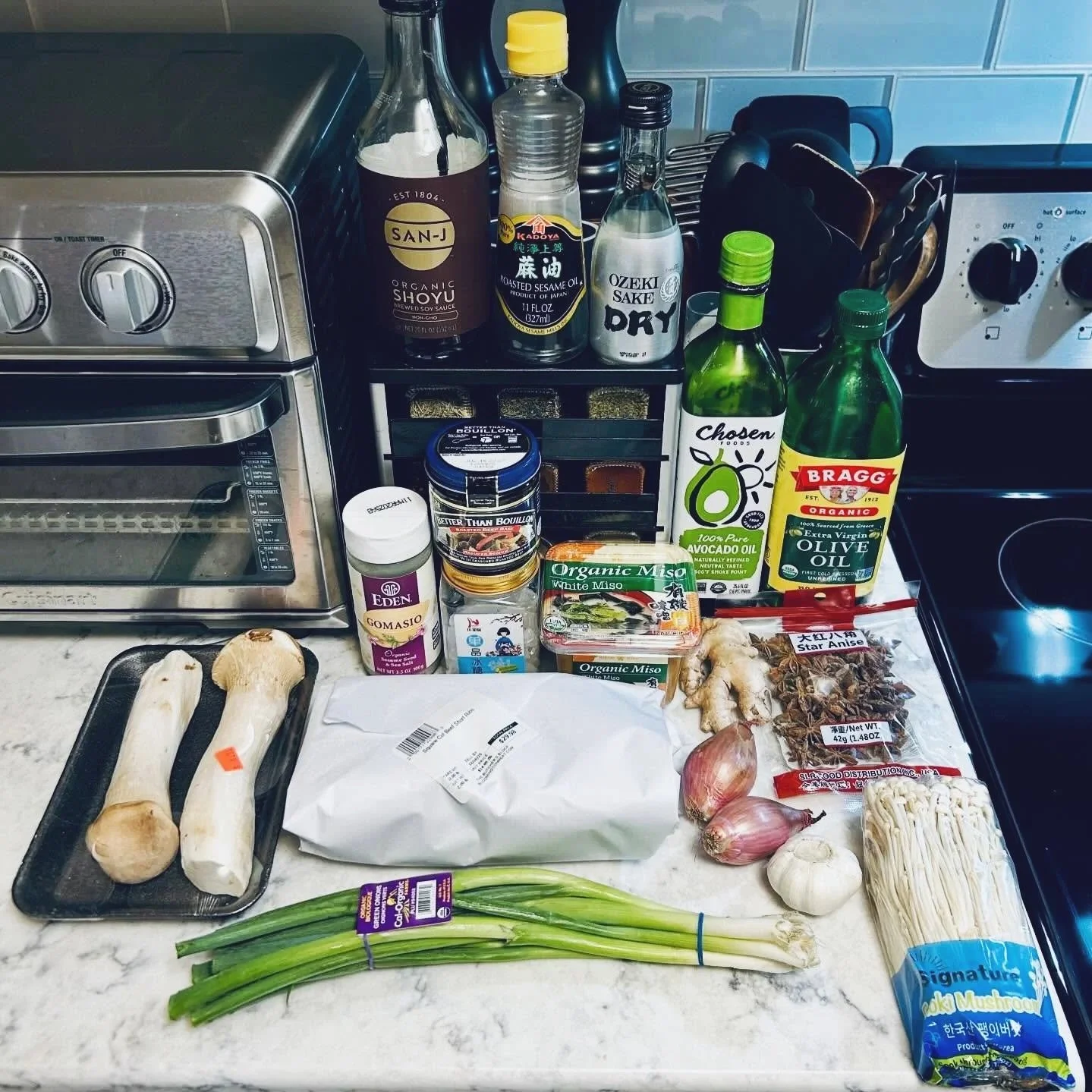 Assorted ingredients and kitchen items on a countertop, including mushrooms, green onions, garlic, ginger, miso paste, soy sauce, sesame oil, olive oil, sake, dried herbs, and a packaged enoki mushroom.