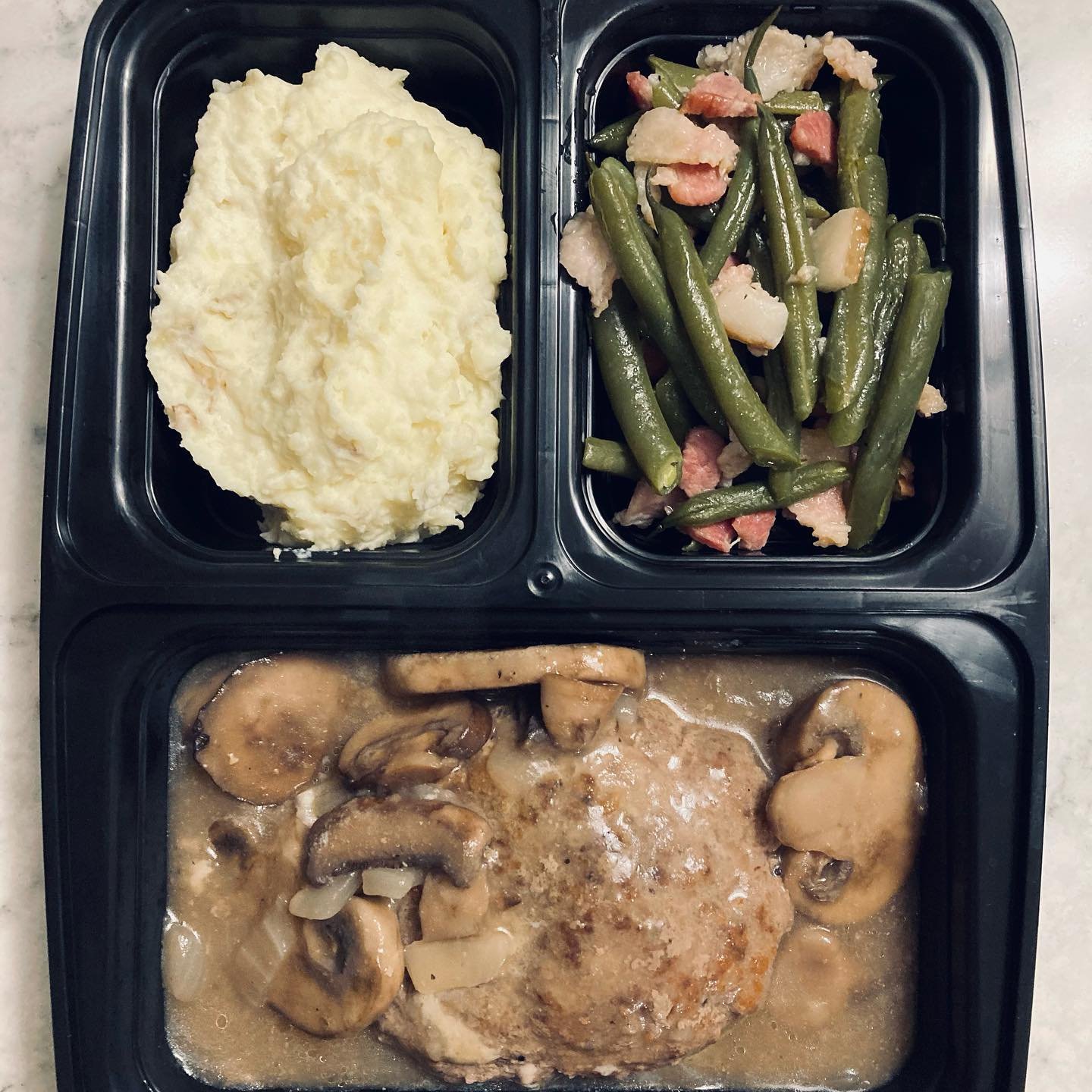 A black tray with three compartments containing mashed potatoes, green beans with bacon, and a chicken cutlet with mushroom gravy.