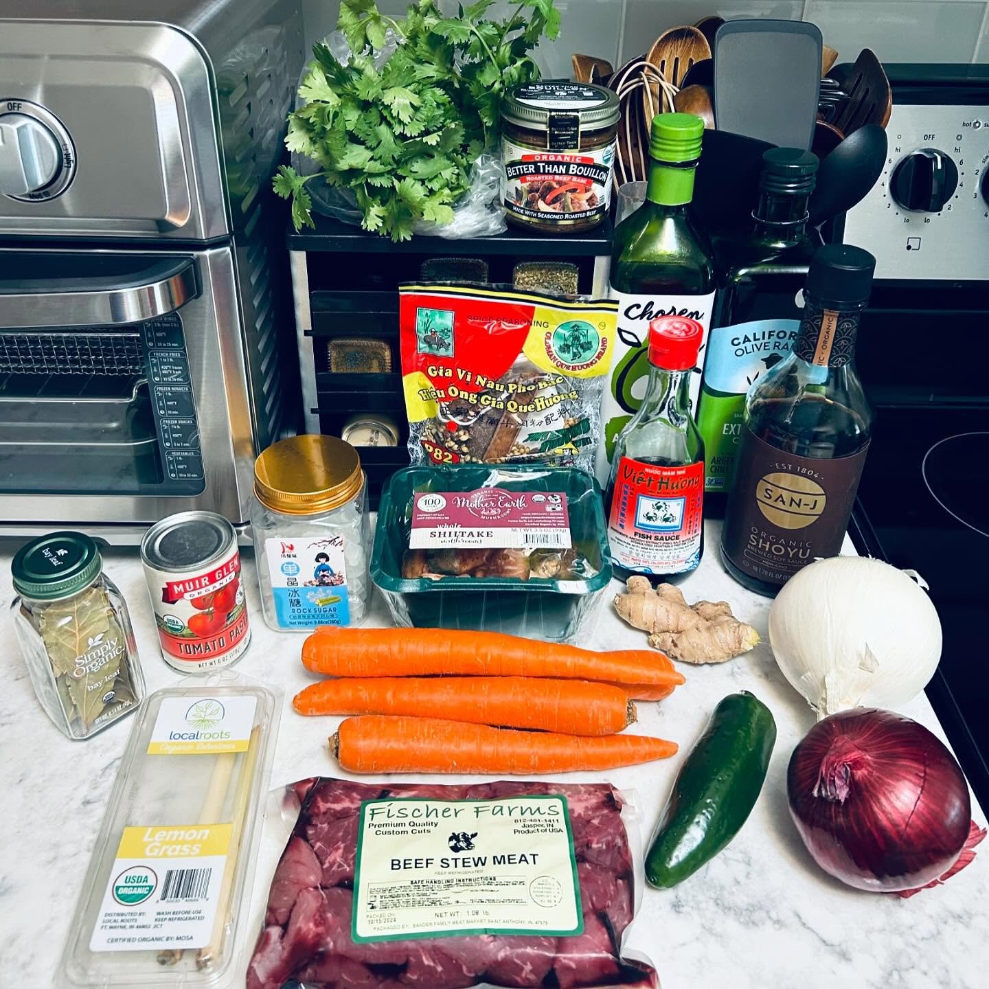 Assorted fresh vegetables, including carrots, cucumber, onion, red onion, and ginger, along with beef stew meat, seasonings, sauces, and herbs on a kitchen countertop.