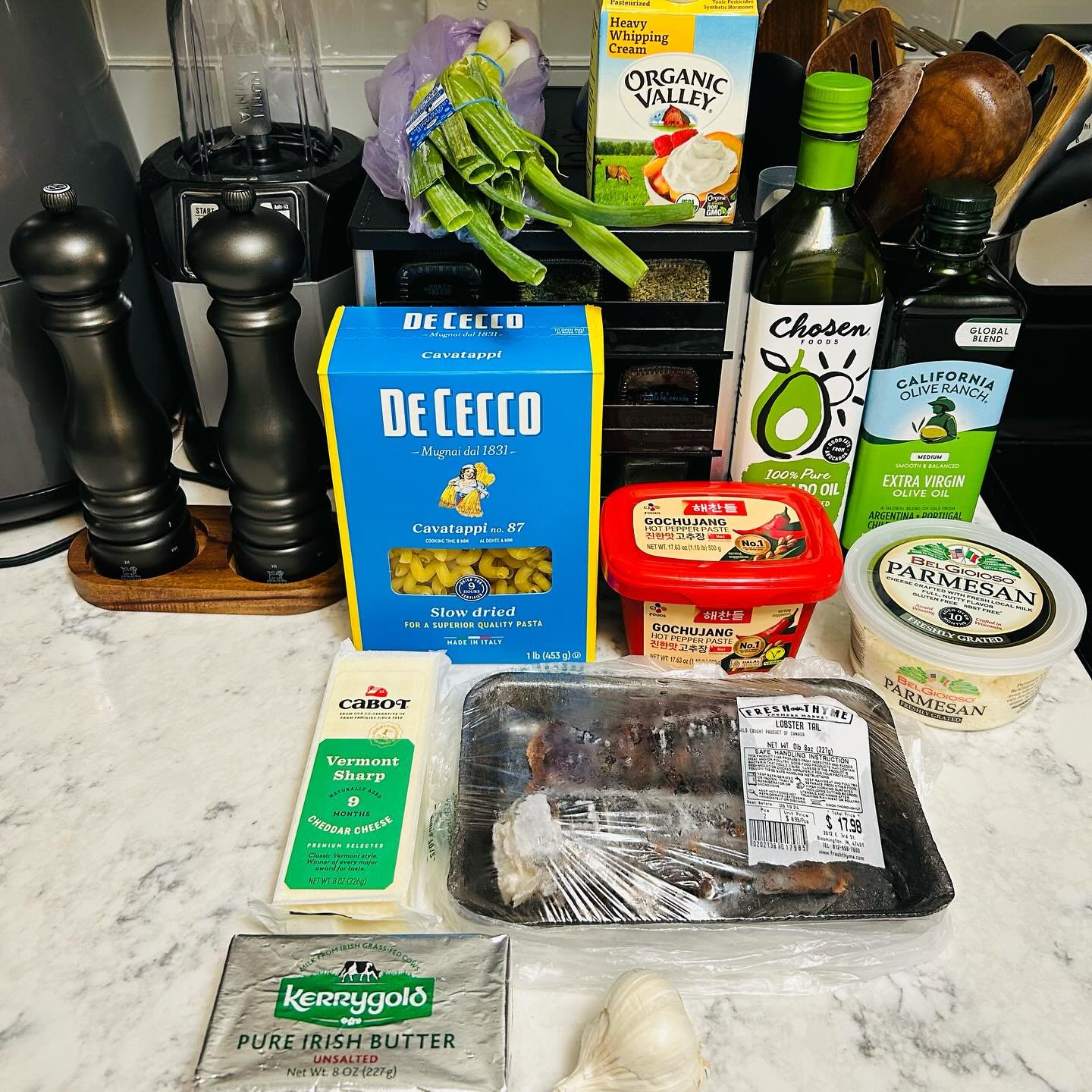 A collection of grocery items on a kitchen countertop, including pasta, cheese, garlic, hot pepper paste, parmesan cheese, a whole raw lobster tail, olive oil bottles, and fresh vegetables like celery and onions, with kitchen appliances and utensils in the background.
