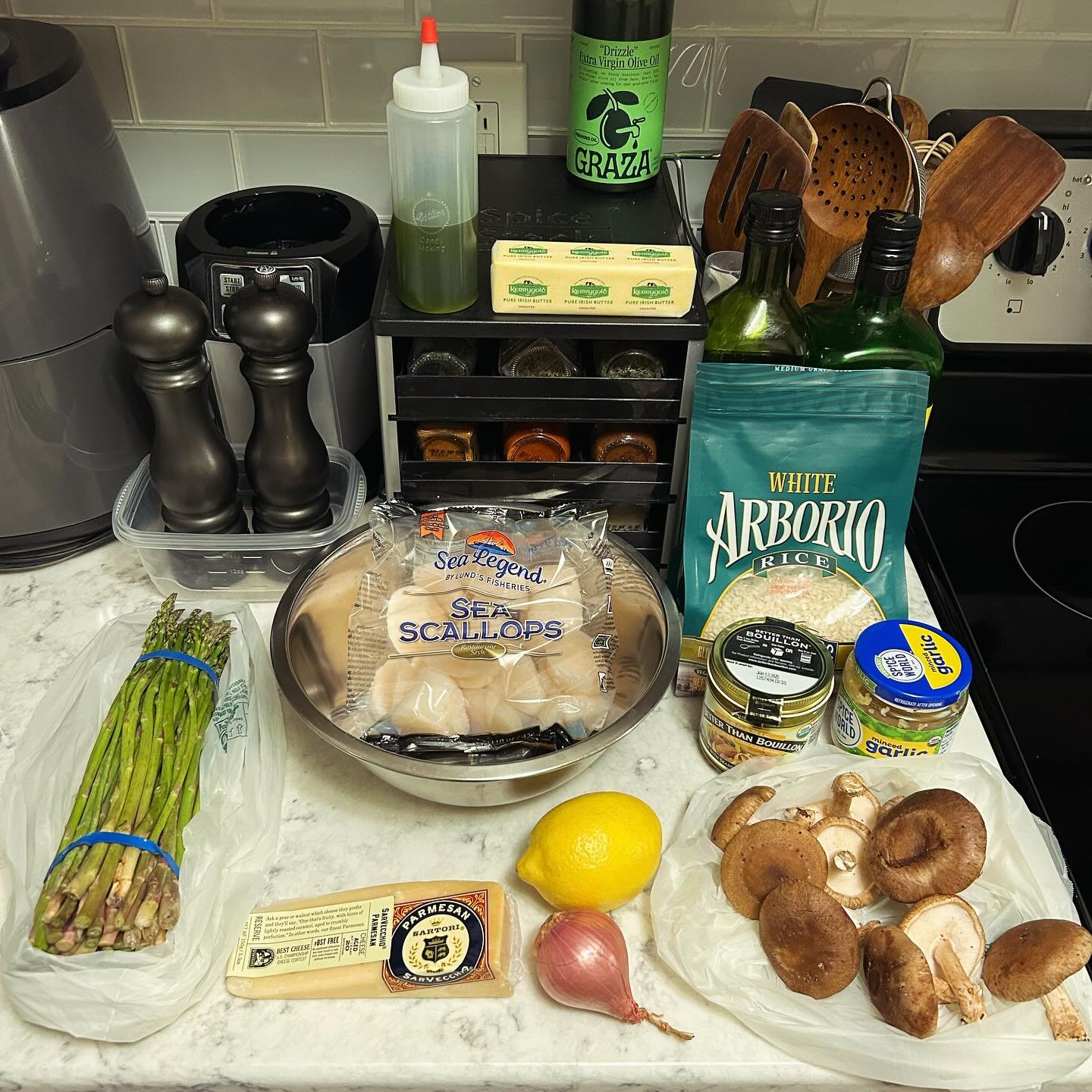 Countertop with fresh ingredients including asparagus, sea scallops, a lemon, shallot, mushrooms, Parmesan cheese, garlic, and bottles of olive oil and seasoning, along with kitchen utensils and condiments.