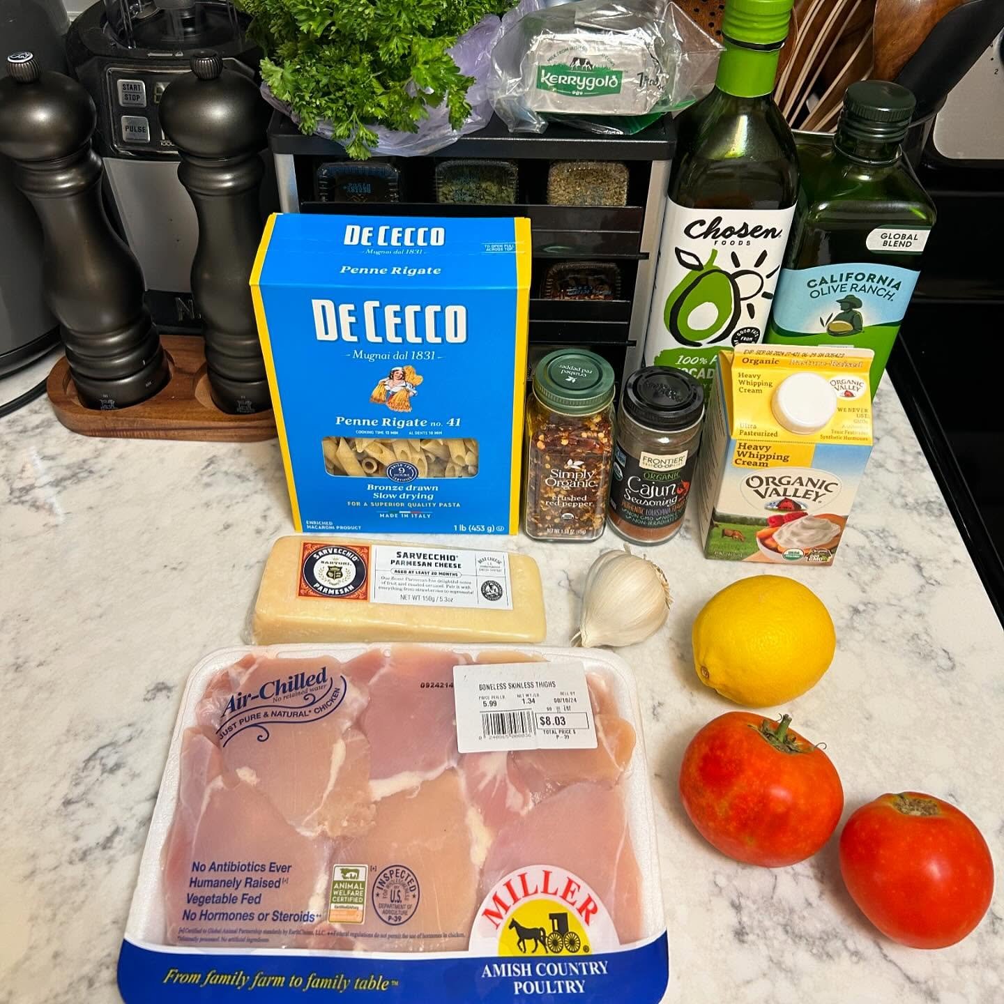 Assorted groceries on a kitchen counter, including pack of chicken, tomatoes, lemon, garlic, pasta, cheese, heavy cream, olive oil, spices, herbs, and pepper grinders.