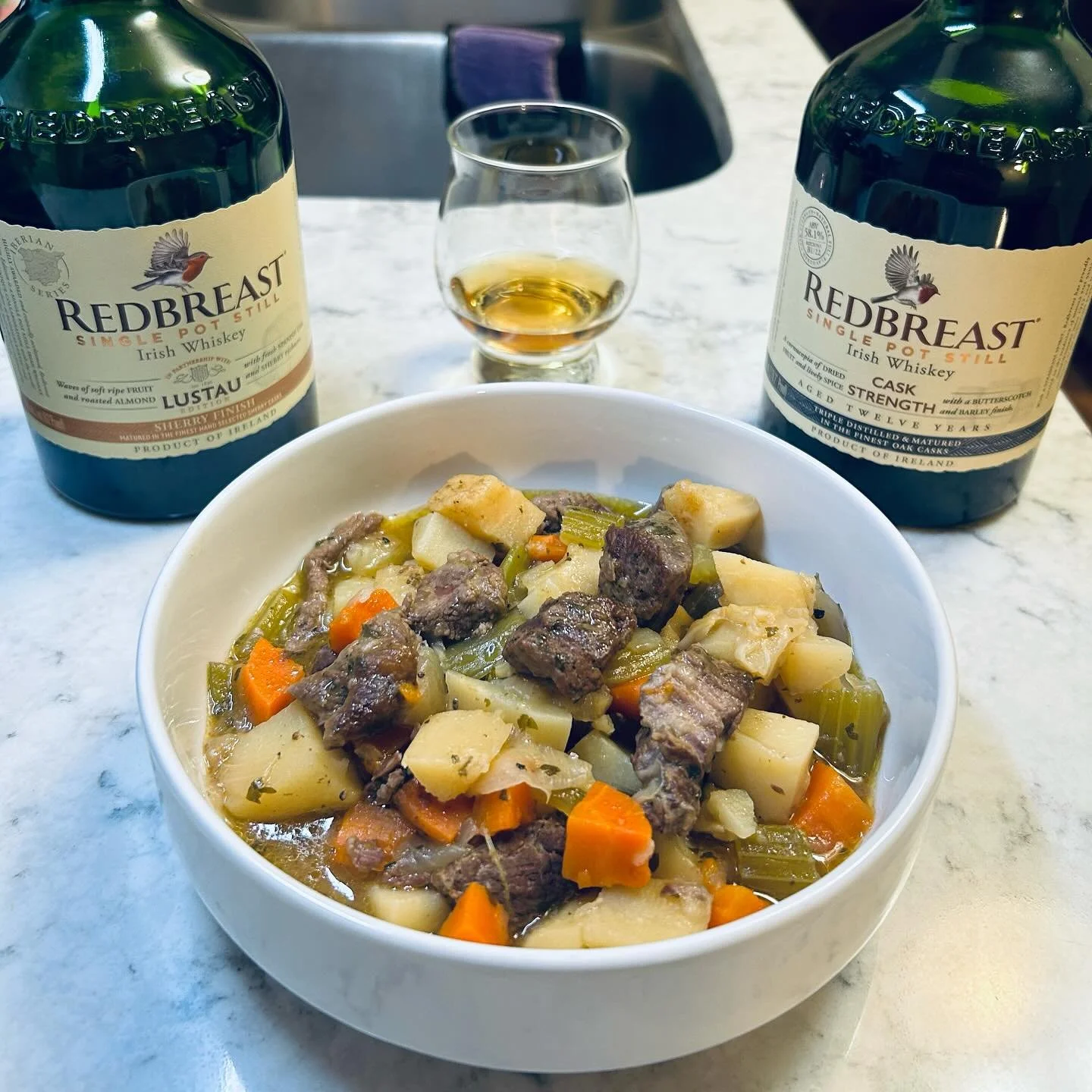 A bowl of beef stew with carrots, potatoes, celery, and chunks of beef on a white marble table, with two bottles of Redbreast Irish whiskey and a glass of whiskey in the background.