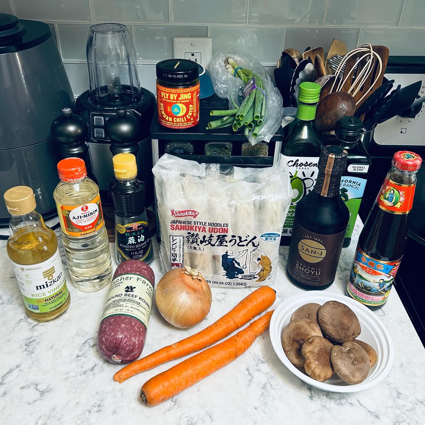A variety of fresh vegetables including carrots, an onion, and mushrooms, along with soy sauce, rice vinegar, and other cooking ingredients on a kitchen countertop.