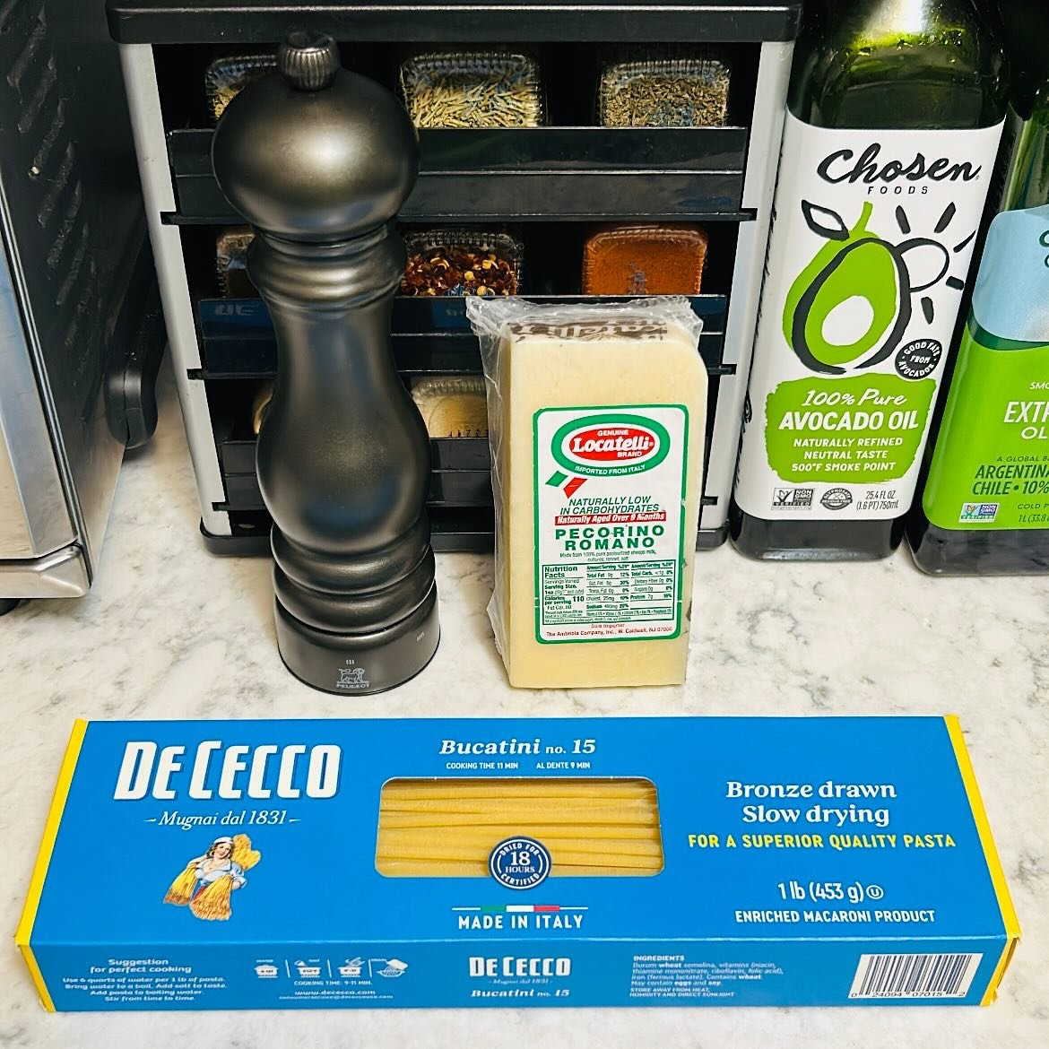 A kitchen counter with a black spice rack containing various spices, a black pepper mill, a package of Pecorino Romano cheese, bottles of avocado oil and other oils, and a box of De Cecco Bucatini pasta.