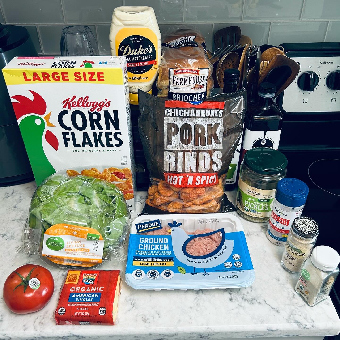 Groceries on a kitchen countertop, including a box of Kellogg's Corn Flakes, a head of lettuce, a tomato, ground chicken, pork rinds, pickles, organic American singles cheese, various spices, mayonnaise, and bread, with kitchen utensils and stove in the background.