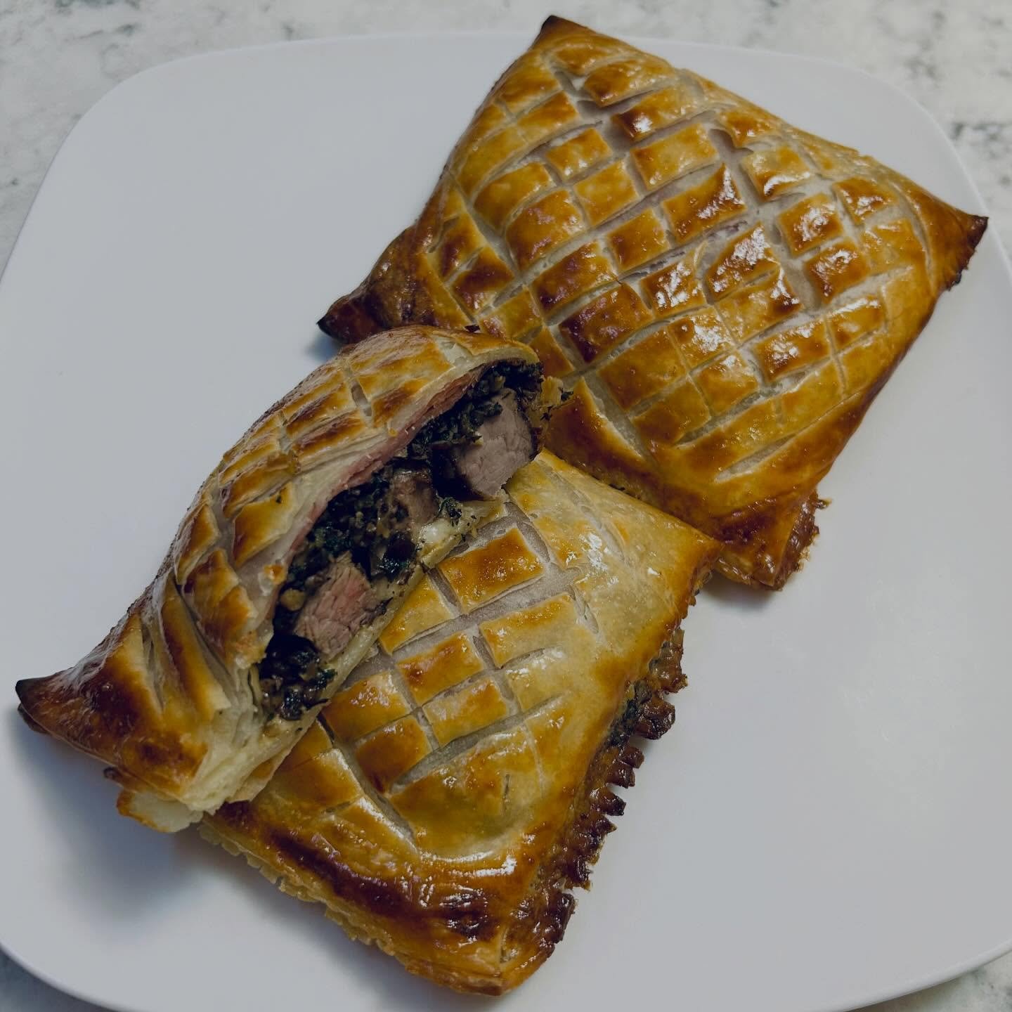 Baked meat Wellington on a white plate, with one piece sliced to show the meat and mushroom filling inside.