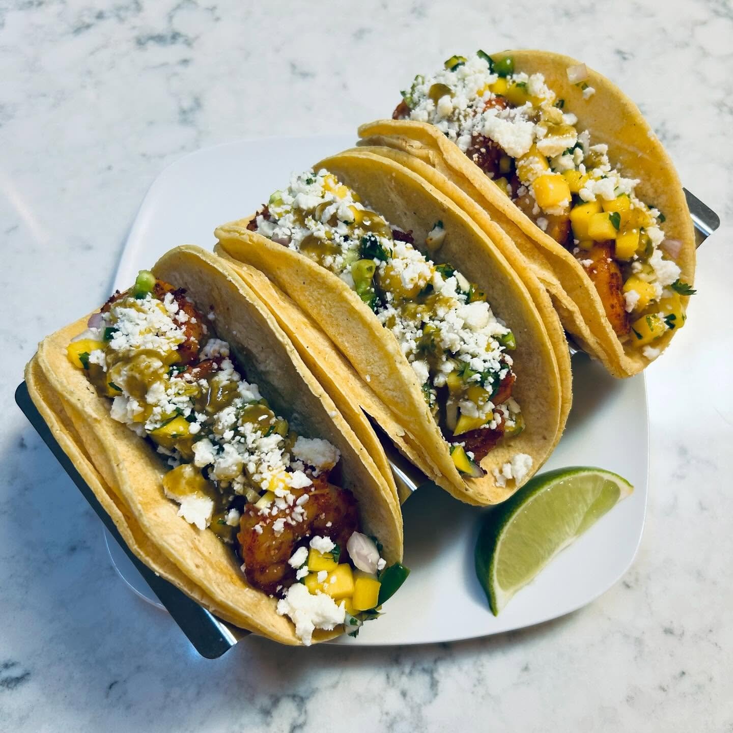 Three tacos filled with chicken, corn, diced onion, shredded cheese, and sauce, served with a lime wedge on a white plate.