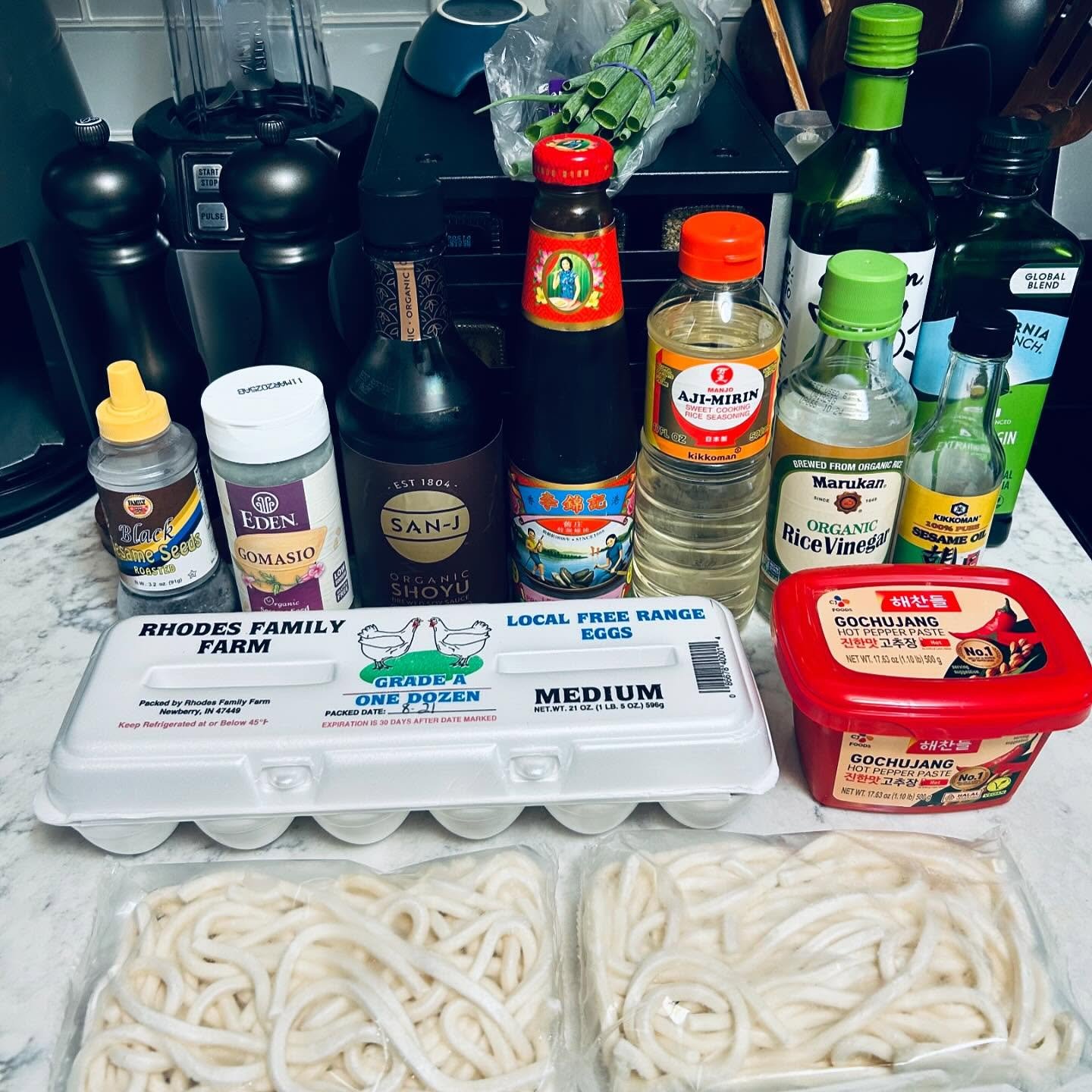A kitchen countertop with various Asian and organic ingredients, including eggs, sesame oil, rice vinegar, soy sauce, hot pepper paste, green onions, and two packs of udon noodles.