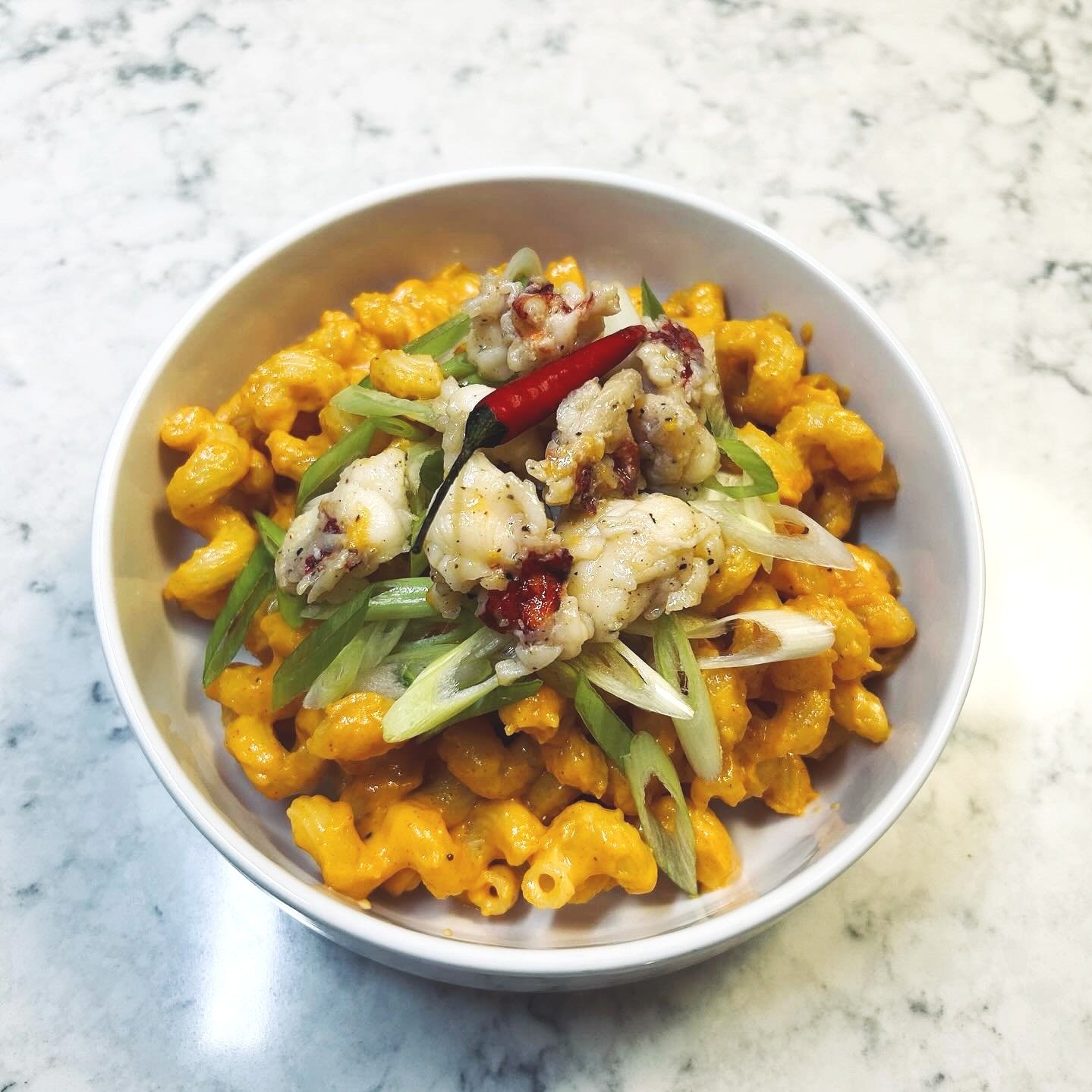 A bowl of macaroni and cheese topped with cooked lobster pieces, sliced green onions, a red chili pepper, and crispy bacon bits on top, placed on a marble surface.