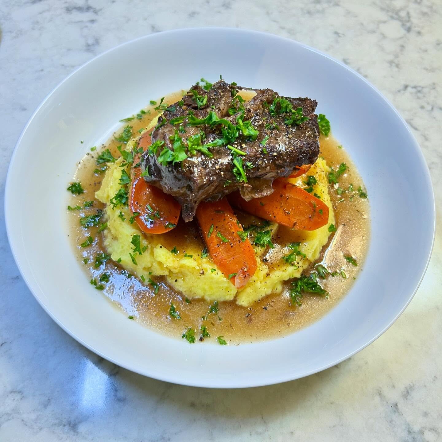 Plate of mashed potatoes topped with a braised beef short rib, cooked carrots, and garnished with chopped parsley, served with gravy.
