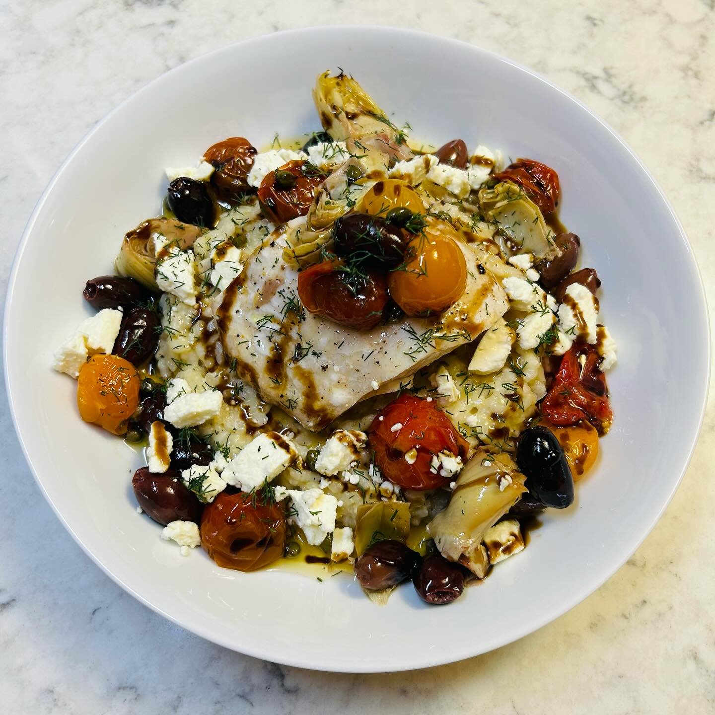 Mediterranean-style dish with cooked fish, cherry tomatoes, olives, crumbled feta cheese, and fresh herbs on a white plate.