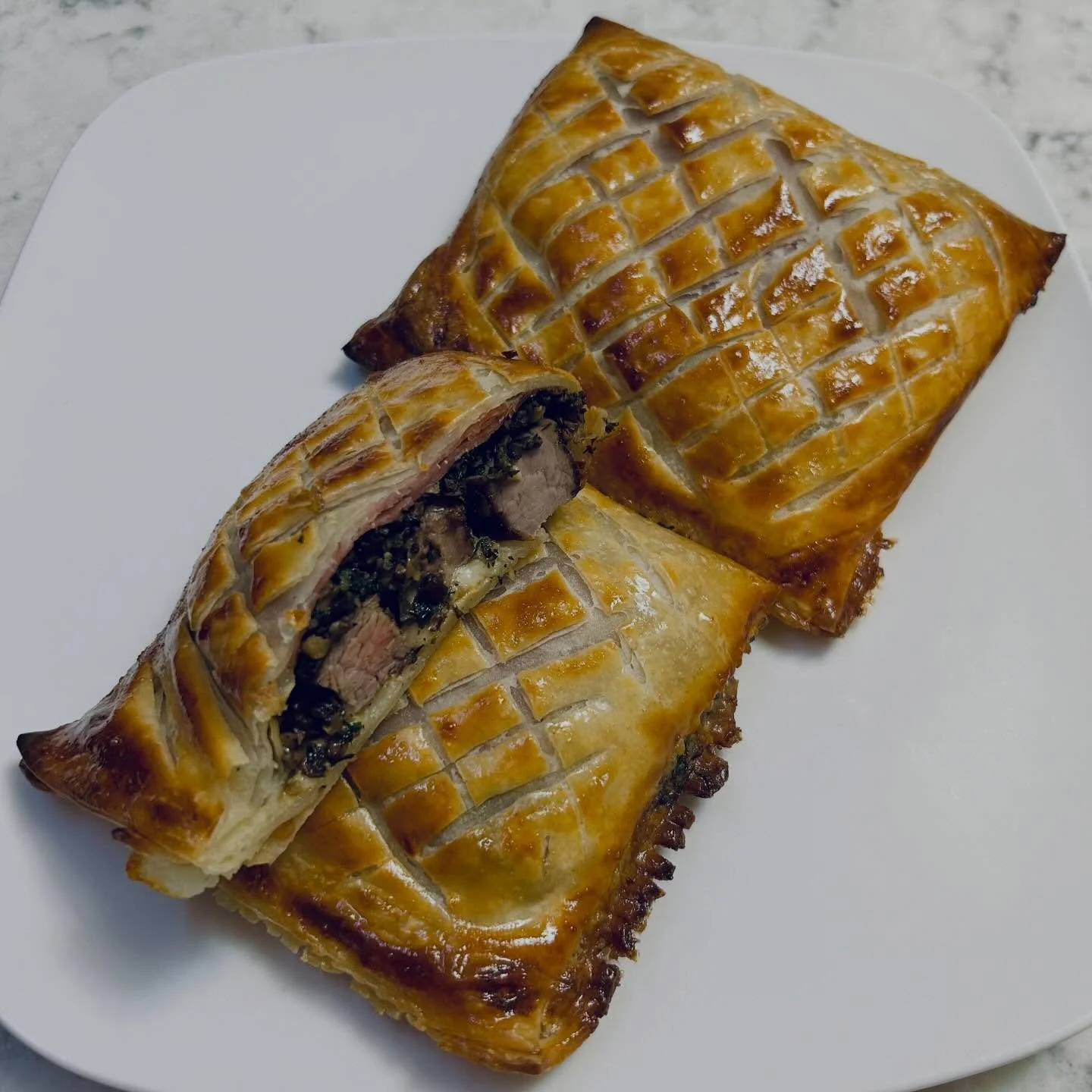 A white plate holding two beef Wellington pastries with golden-brown, scored pastry crusts, one cut open showing beef, mushroom duxelles, and puff pastry filling.