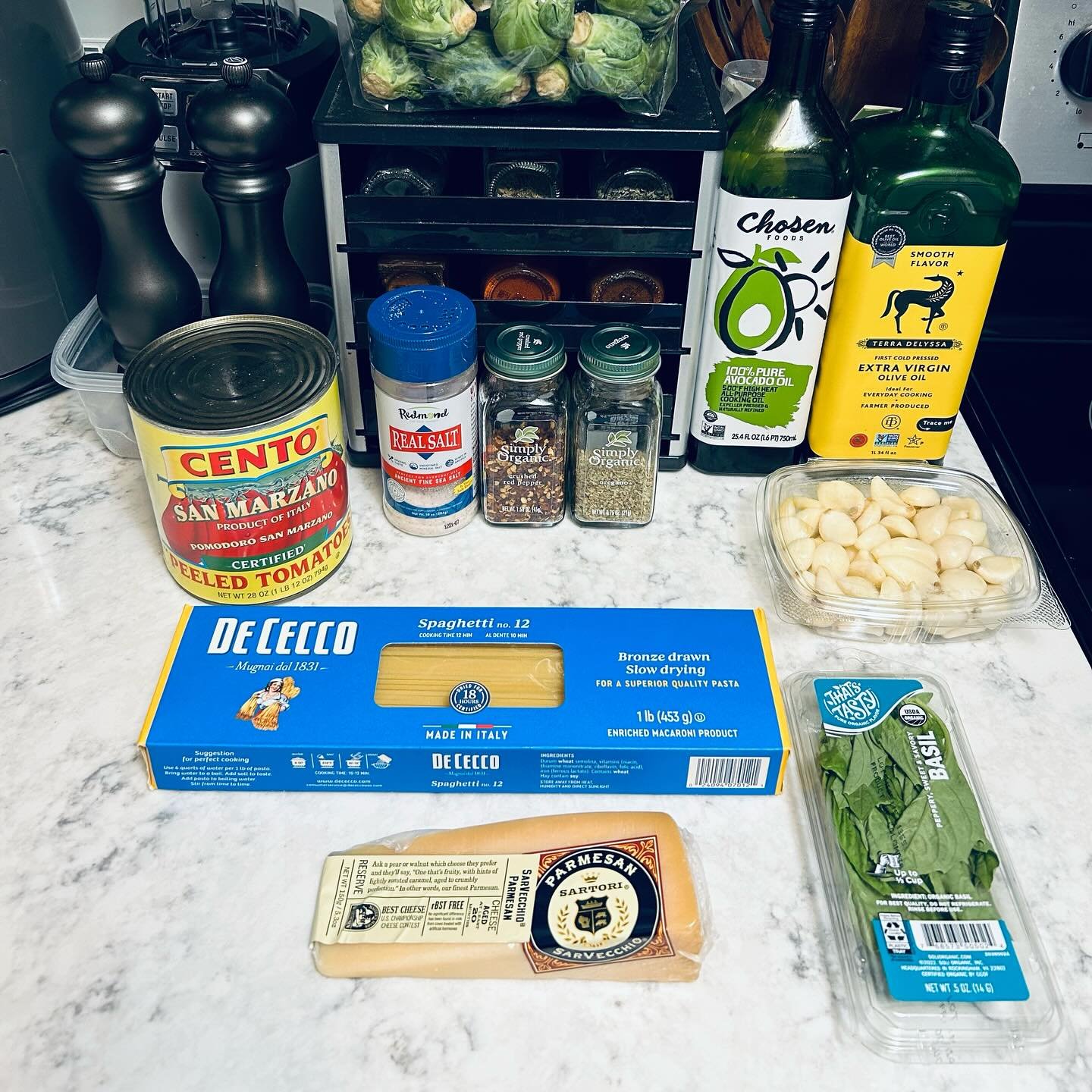 Spaghetti pasta, fresh basil, Parmesan cheese, canned peeled tomatoes, garlic cloves, olive oils, salt, pepper, and Italian seasonings on a kitchen countertop.
