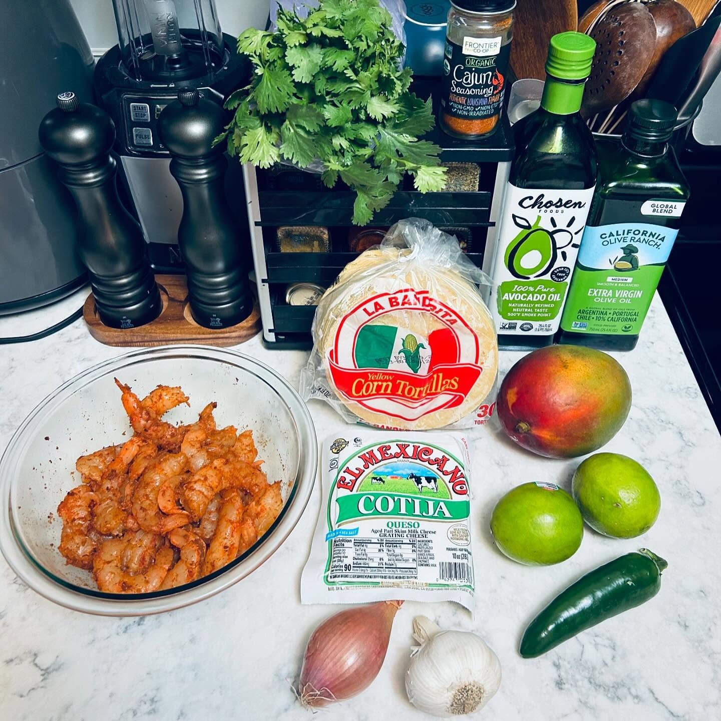 Raw shrimp in a glass bowl, a bag of corn tortillas, queso cheese, a mango, two limes, a jalapeño pepper, garlic, a shallot, cilantro, cooking oil, seasonings, and various kitchen utensils on a white countertop.