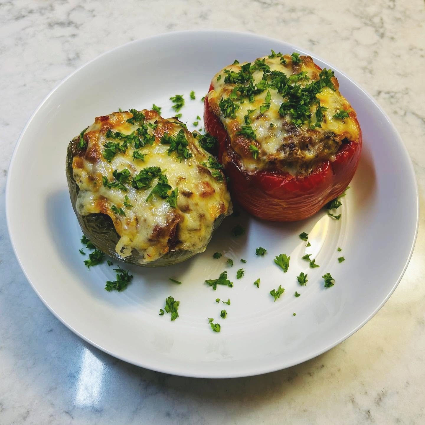 Two stuffed bell peppers, one green and one red, topped with melted cheese and chopped herbs, on a white plate.