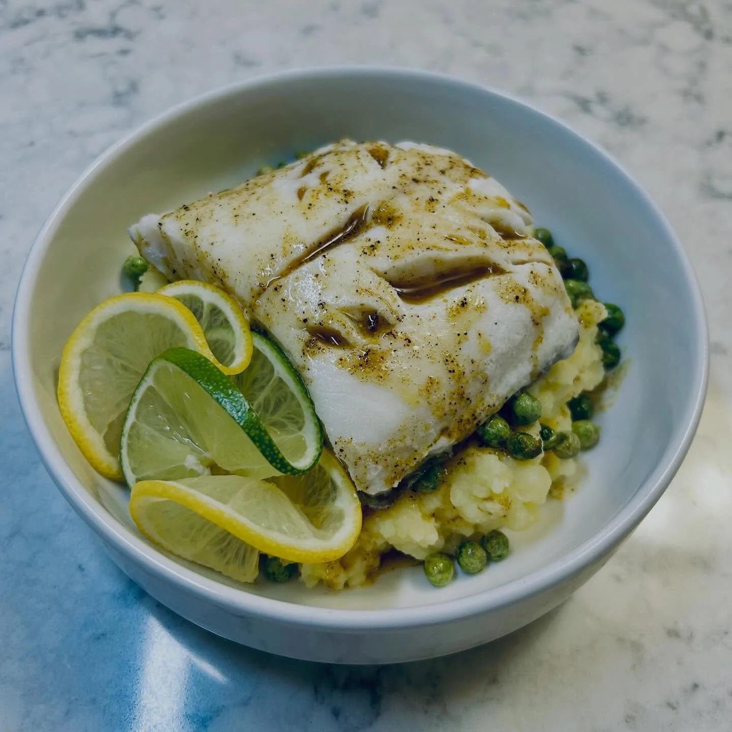 A bowl of cooked white fish with lemon and lime slices, green peas, and cauliflower, garnished with herbs and spices.
