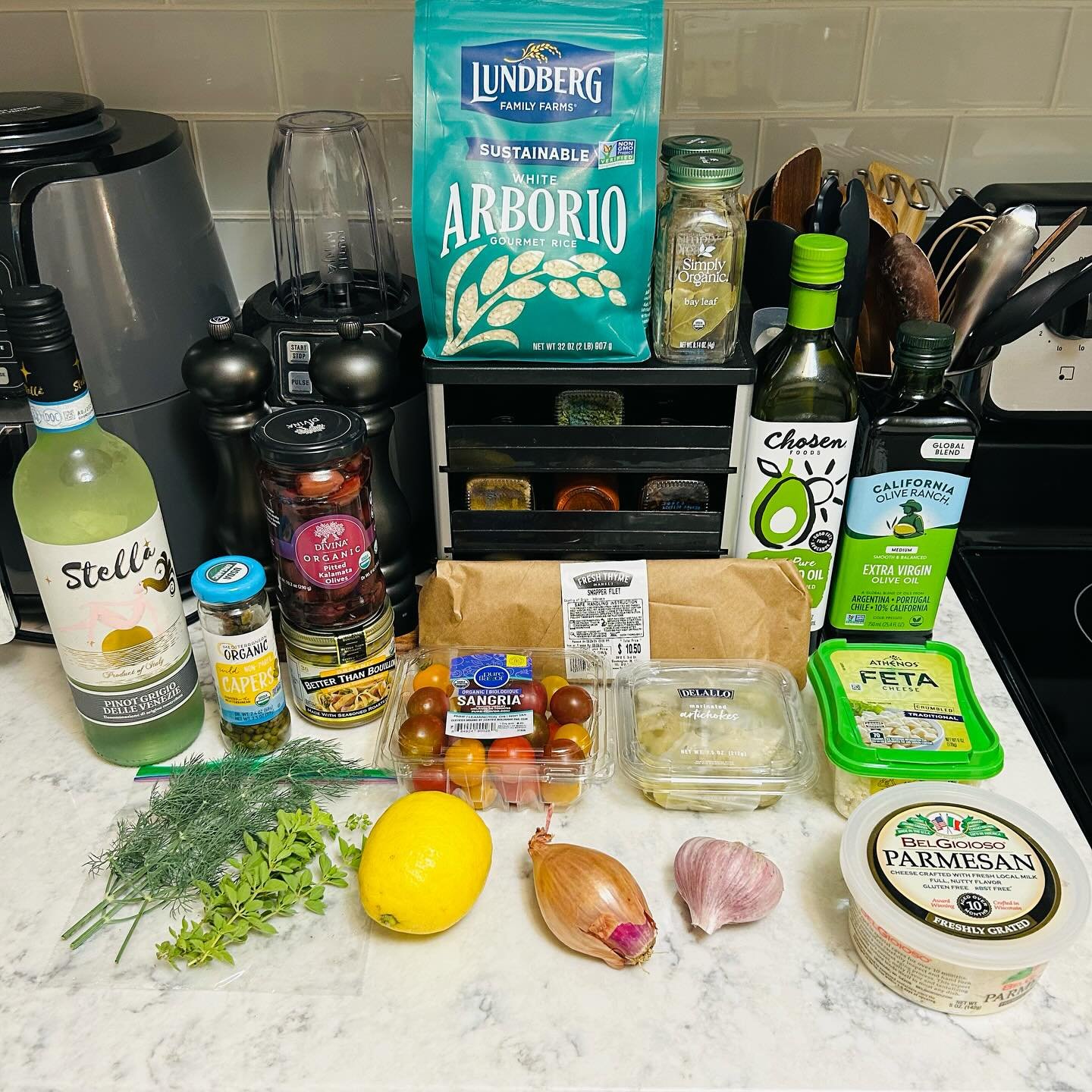 Assorted ingredients on a kitchen counter, including a bottle of prickly pear flavored drink, a jar of capers, cherry tomatoes, a lemon, garlic, fresh herbs, artichokes, parmesan cheese, and olive oil.