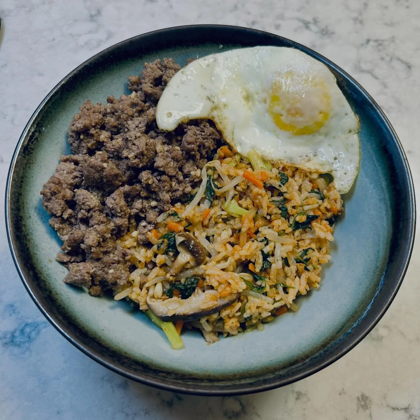 A bowl of ground beef, fried egg, and fried rice with vegetables on a gray marble surface.