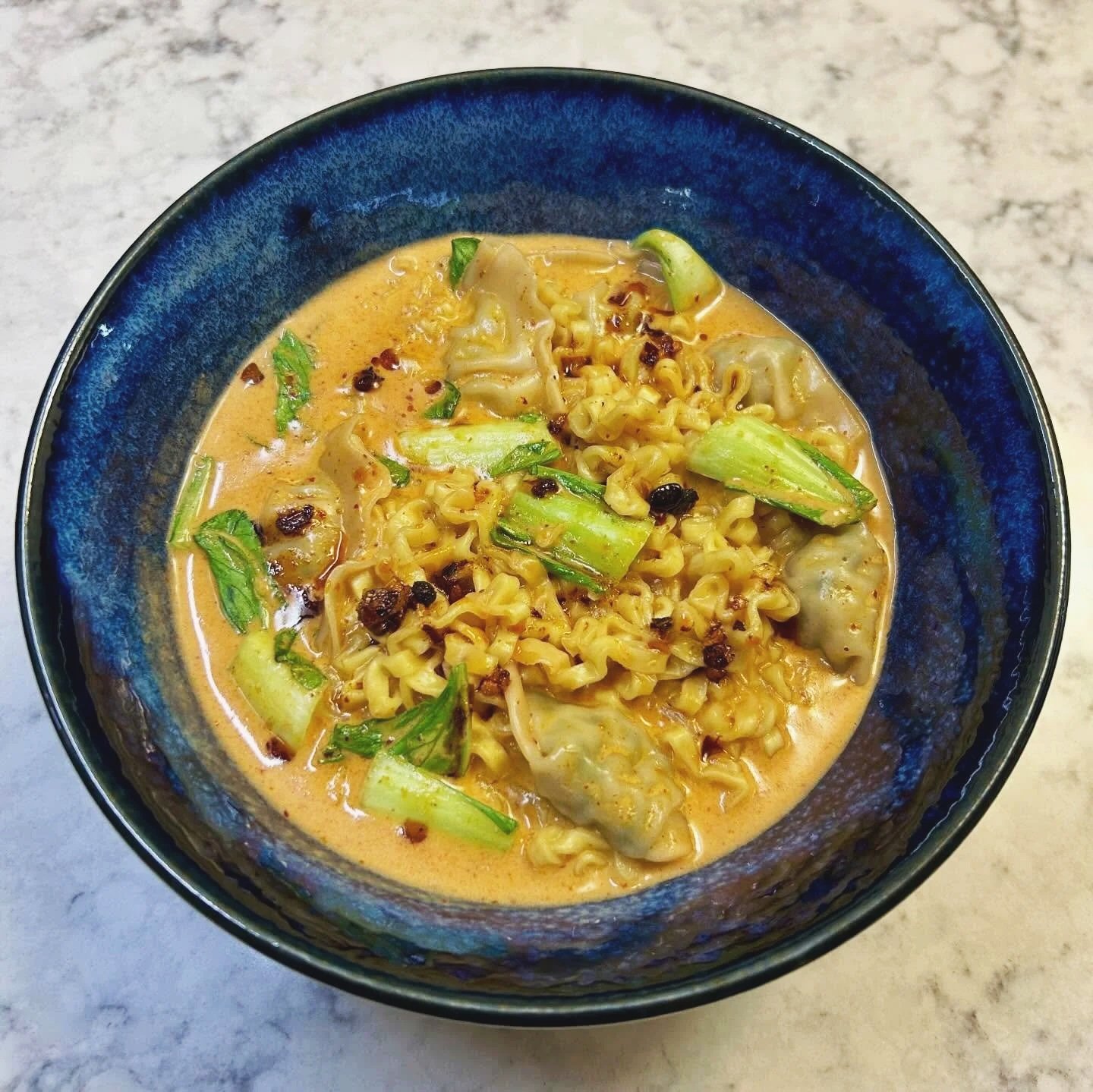A bowl of ramen noodles with vegetables in a creamy broth, garnished with chopped green onions and red pepper flakes