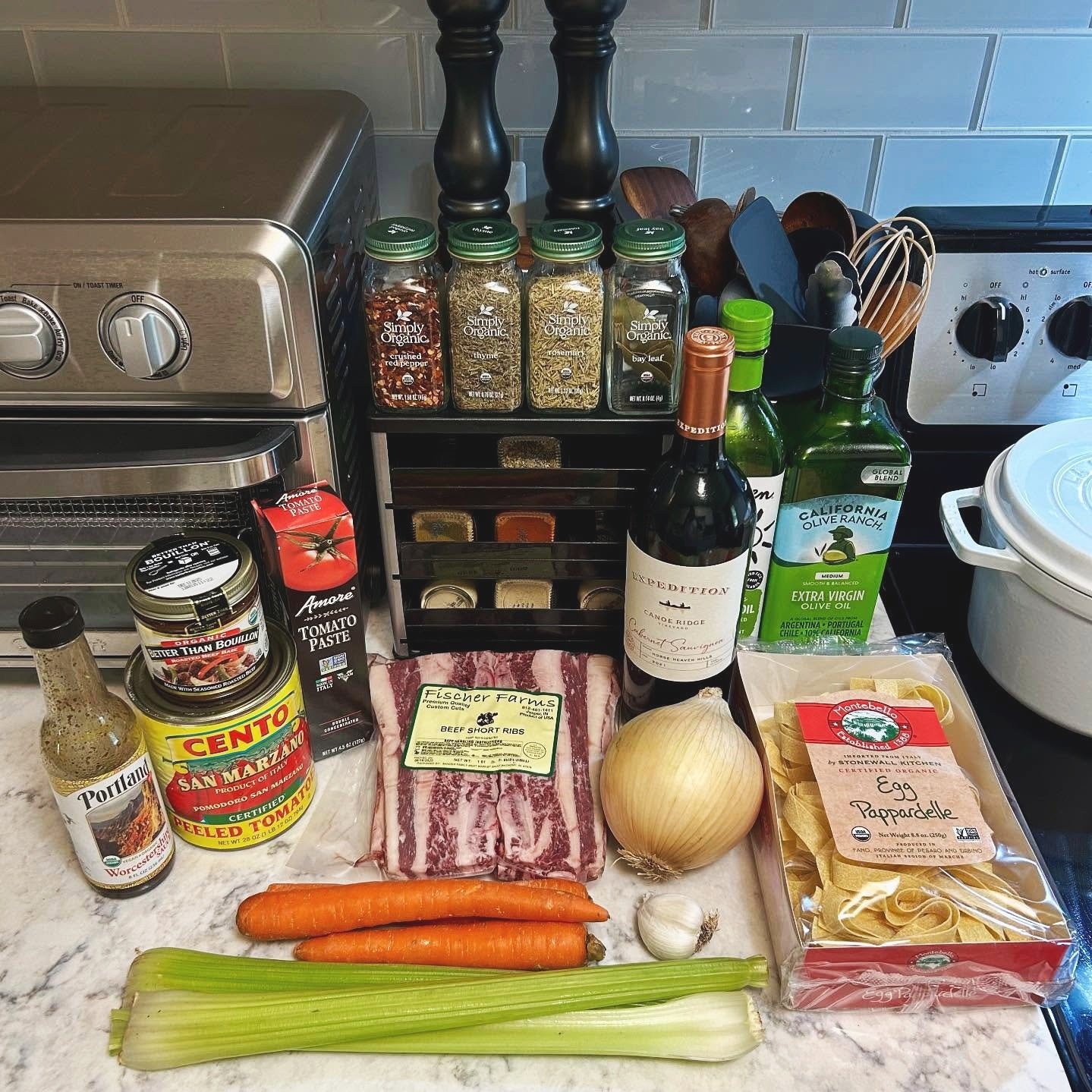 A countertop with various cooking ingredients and supplies including beef short ribs, carrots, celery, onion, garlic, tomato paste, canned tomatoes, spices, olive oil, and pasta, with a spice rack, toaster oven, and containers in the background.