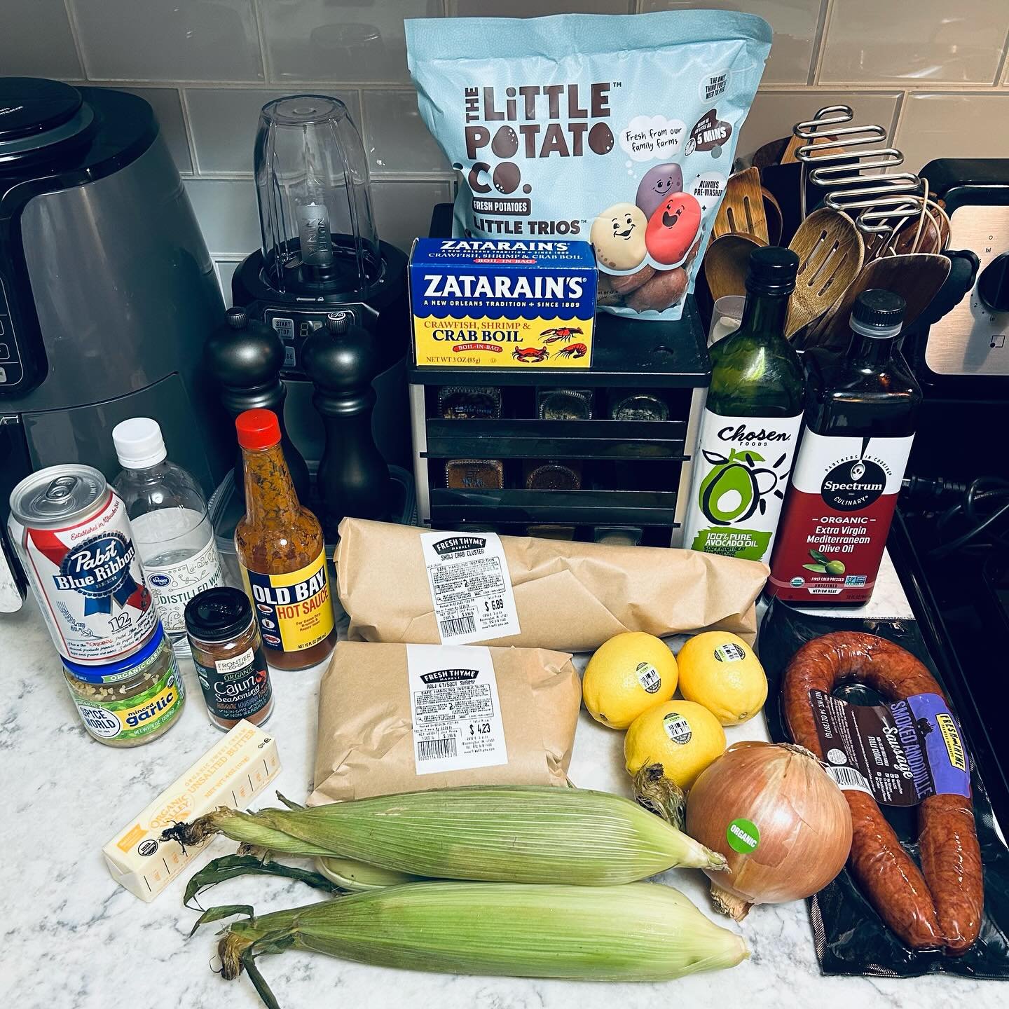 Grocery items on a kitchen counter including corn, onion, lemons, sausage, sauces, spices, and packaged foods with a toaster and utensils in the background.