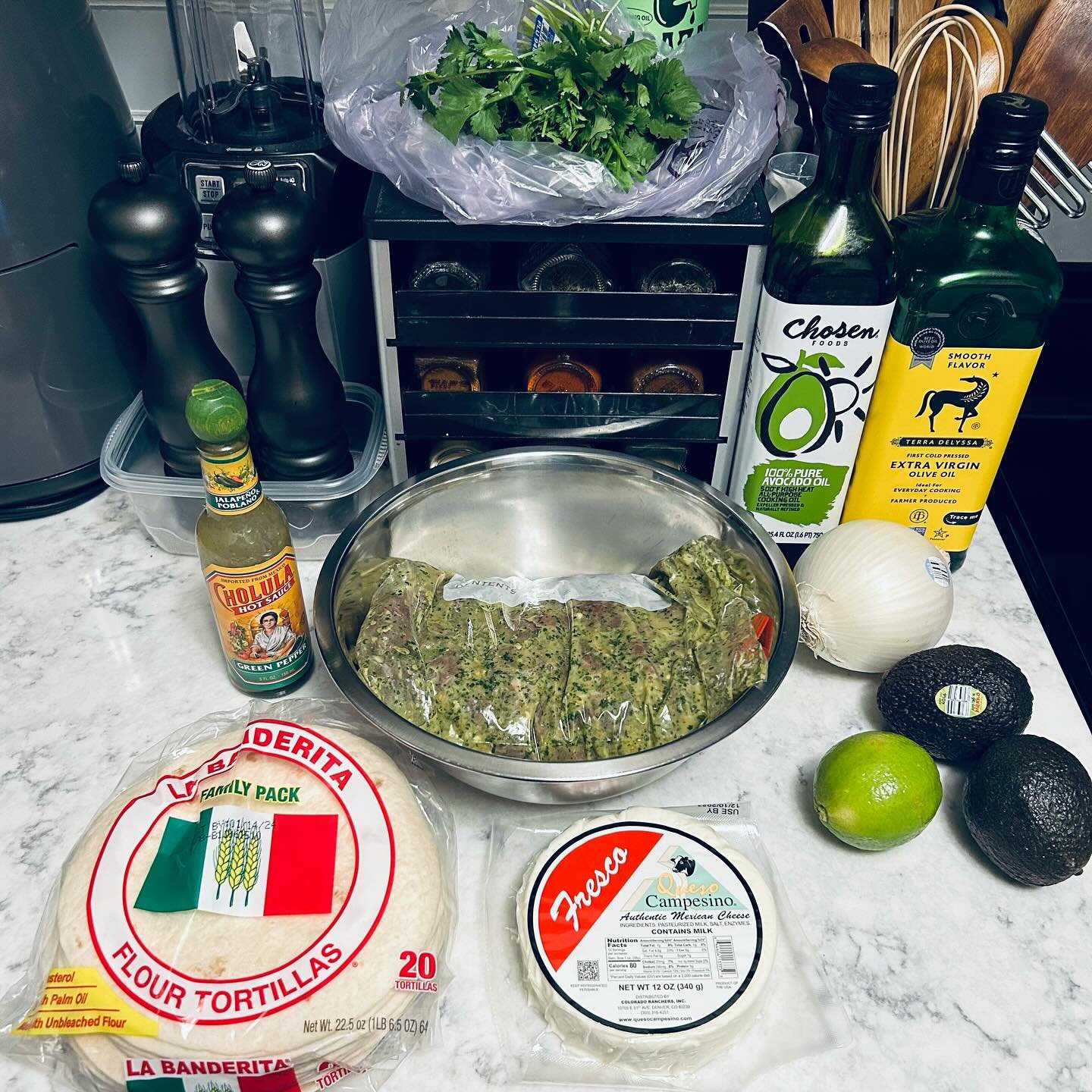 Ingredients laid out on a kitchen countertop, including flour tortillas, queso cheese, avocados, limes, onion, hot sauce, olive oil, cilantro, and a large marinated meat piece, with spices, kitchen utensils, and appliances in the background.