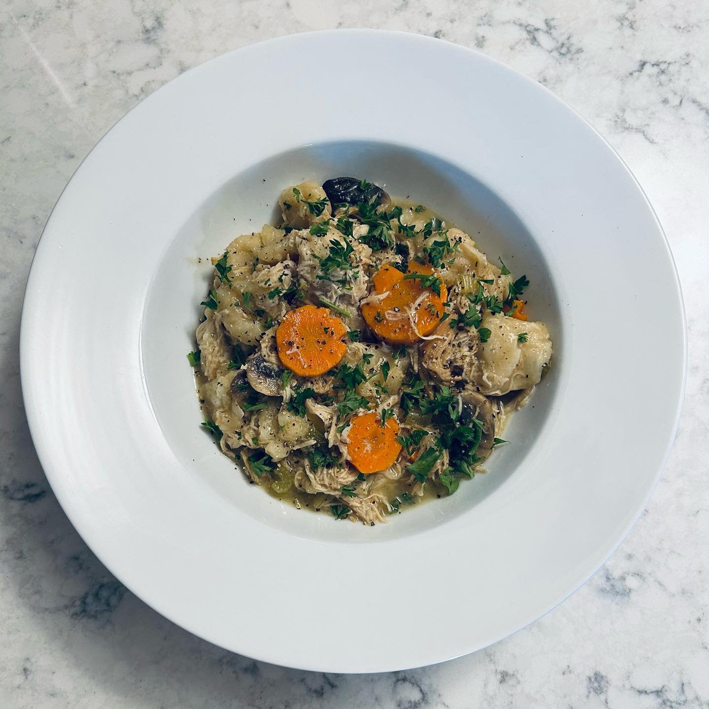 A white plate of chicken noodle soup with carrots, mushrooms, herbs, and shredded chicken, served on a marble surface.