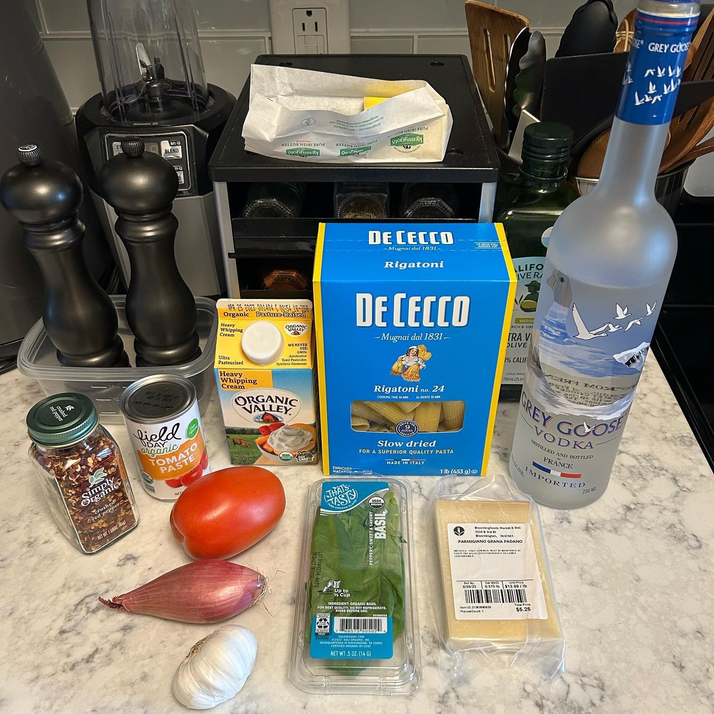Ingredients for a pasta dish including basil, onion, garlic, tomato, grated Parmigiano cheese, heavy whipping cream, organic tomato paste, a box of De Cecco rigatoni pasta, and a bottle of Grey Goose vodka.