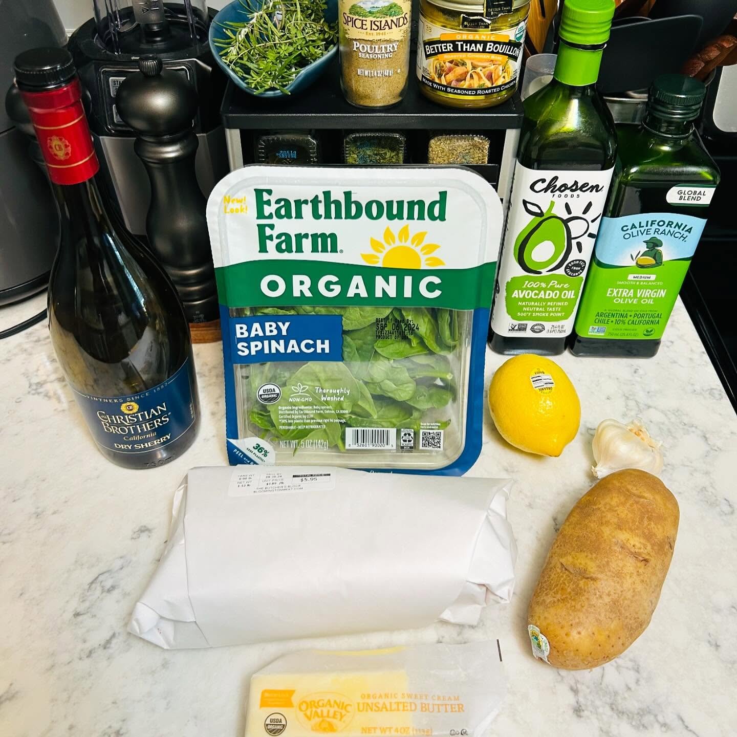A selection of groceries on a kitchen countertop including a bottle of Christian Brothers Dry Sherry, a container of Earthbound Farm organic baby spinach, a lemon, a sweet potato, a head of garlic, a stick of unsalted butter, and bottles of avocado oil, olive oil, and seasoning among other items.