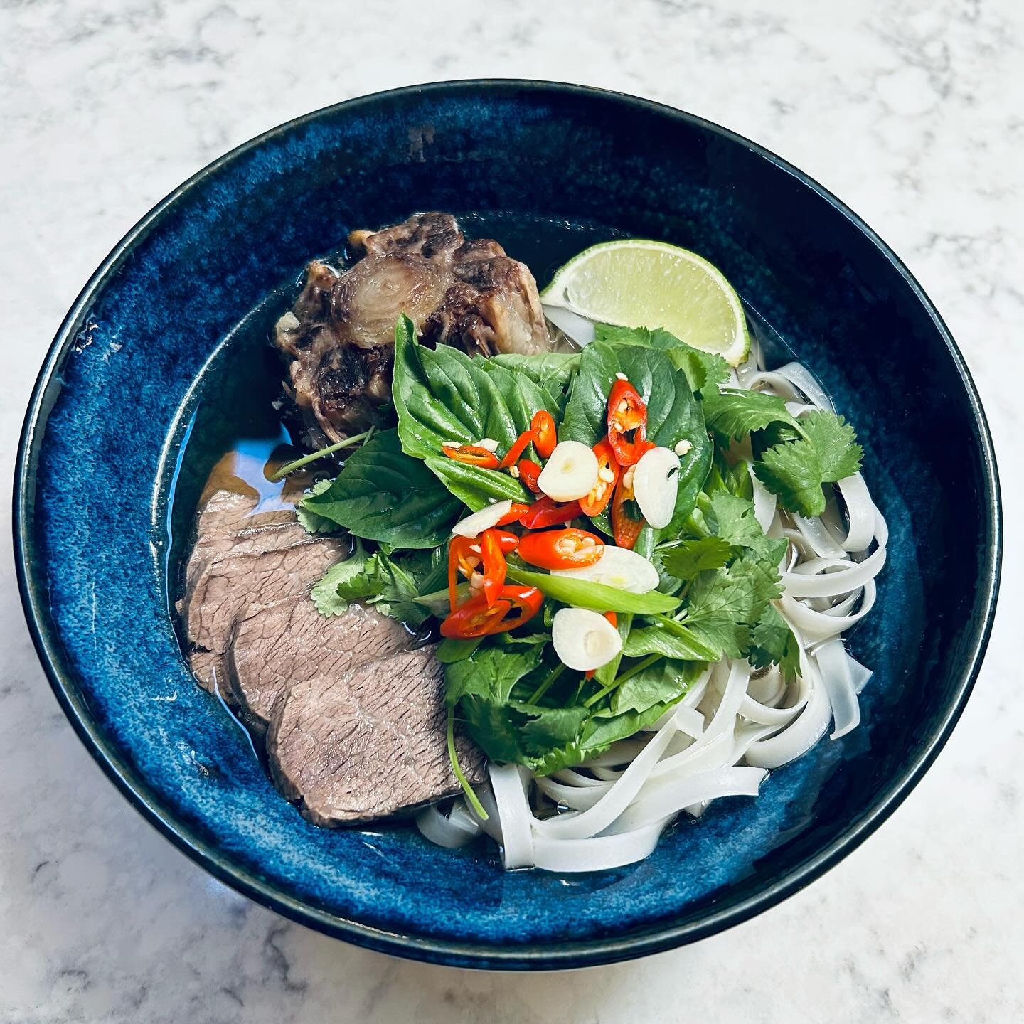 A bowl of Vietnamese pho with rice noodles, slices of beef, chopped herbs, sliced chili peppers, garlic, a lime wedge, and a piece of beef marrow bone, on a light-colored marble surface.