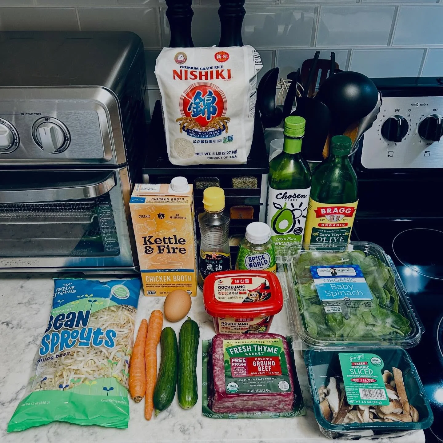 Assorted ingredients and kitchen items on a countertop, including a bag of Nishiki rice, a carton of chicken broth, bean sprouts, carrots, cucumbers, an egg, fresh thyme, sliced mushrooms, and condiments like soy sauce and hot pepper paste.