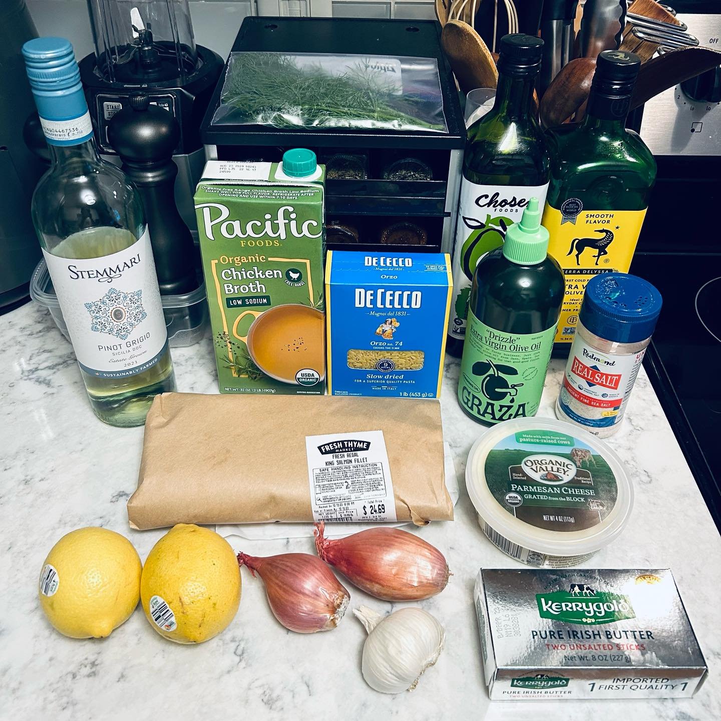 Groceries on a kitchen counter including lemons, garlic, shallots, butter, chicken, pasta, cheese, olive oil, salt, and beverages. In the background, there are kitchen utensils and appliances.
