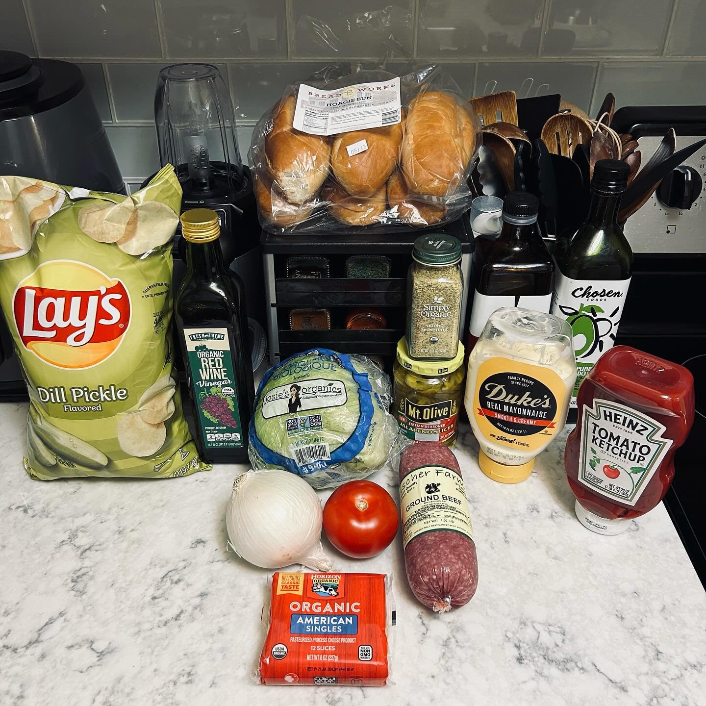 Assorted grocery items including Lay's dill pickle flavored chips, organic red wine vinegar, a head of cauliflower, a white onion, a tomato, ground beef, sliced cheese, a loaf of bread, mayonnaise, tomato ketchup, hot sauce, a jar of green olives, a jar of jalapeño slices, and bottles of balsamic vinegar and other condiments on a kitchen countertop.