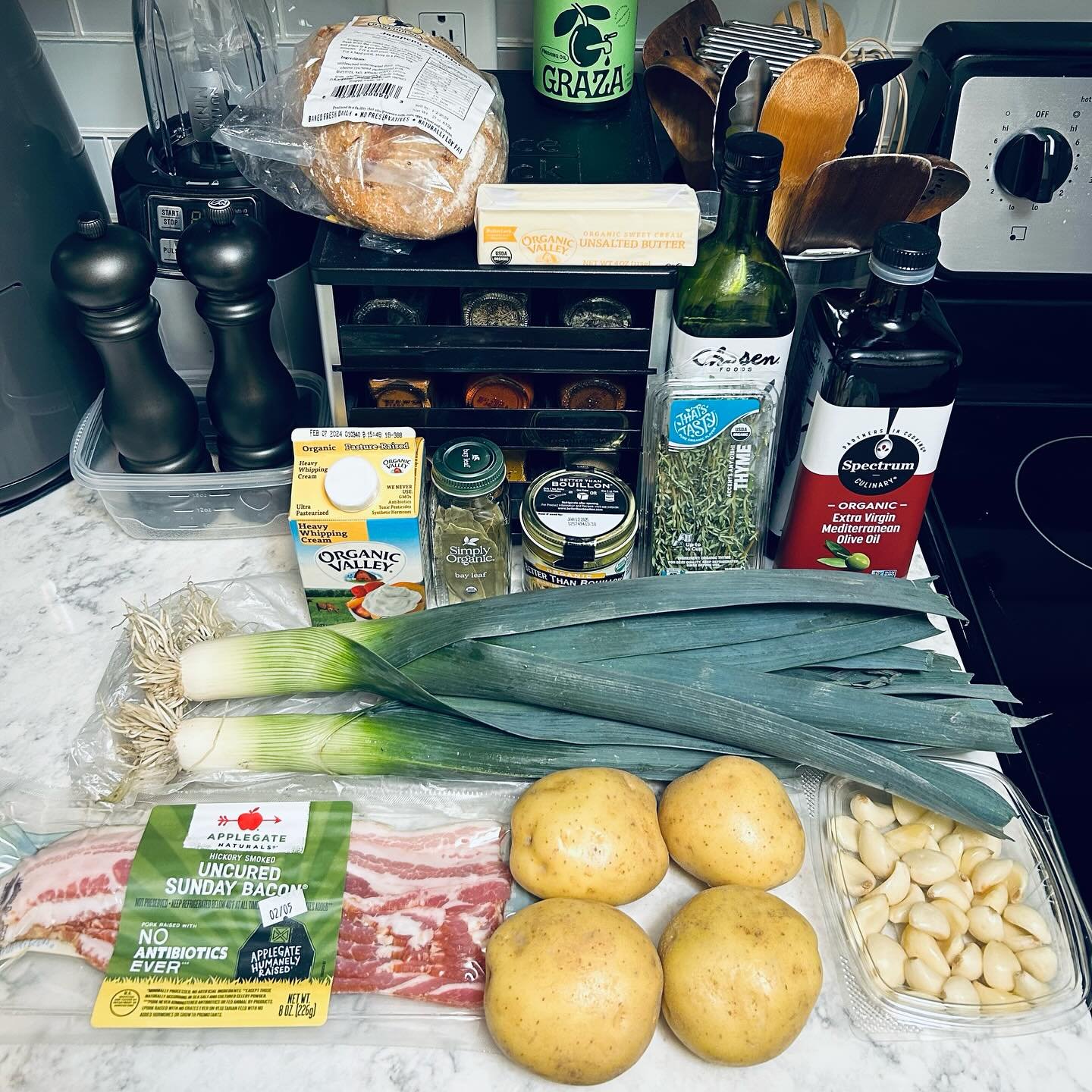 Fresh vegetables such as potatoes, garlic cloves, and green onions on a kitchen counter with various herbs, spices, olive oil, bacon, bread, and condiments behind them.