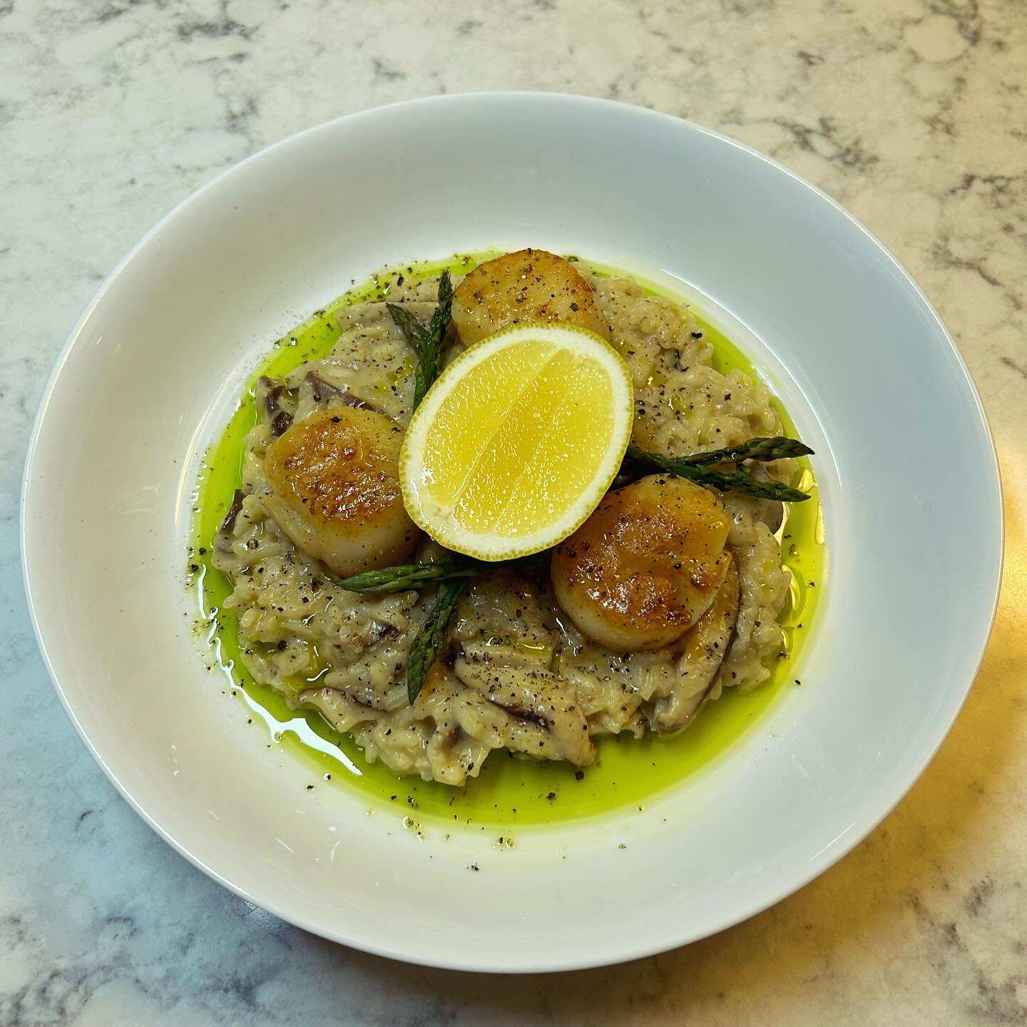 A white bowl of creamy risotto topped with seared scallops, grilled asparagus, lemon wedge, black pepper, and olive oil.