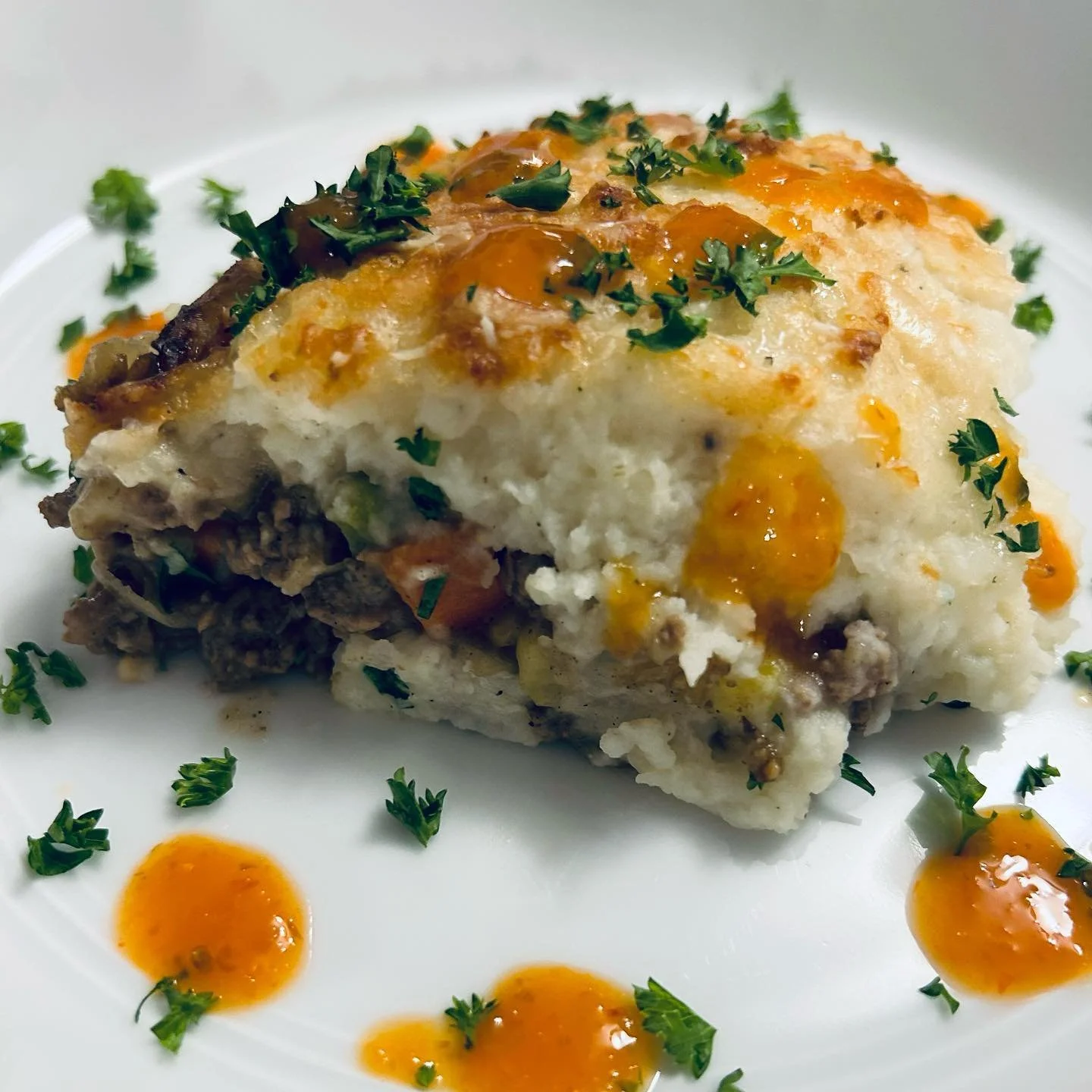 A serving of shepherd's pie topped with mashed potatoes, garnished with chopped parsley and drizzled with orange sauce, on a white plate.