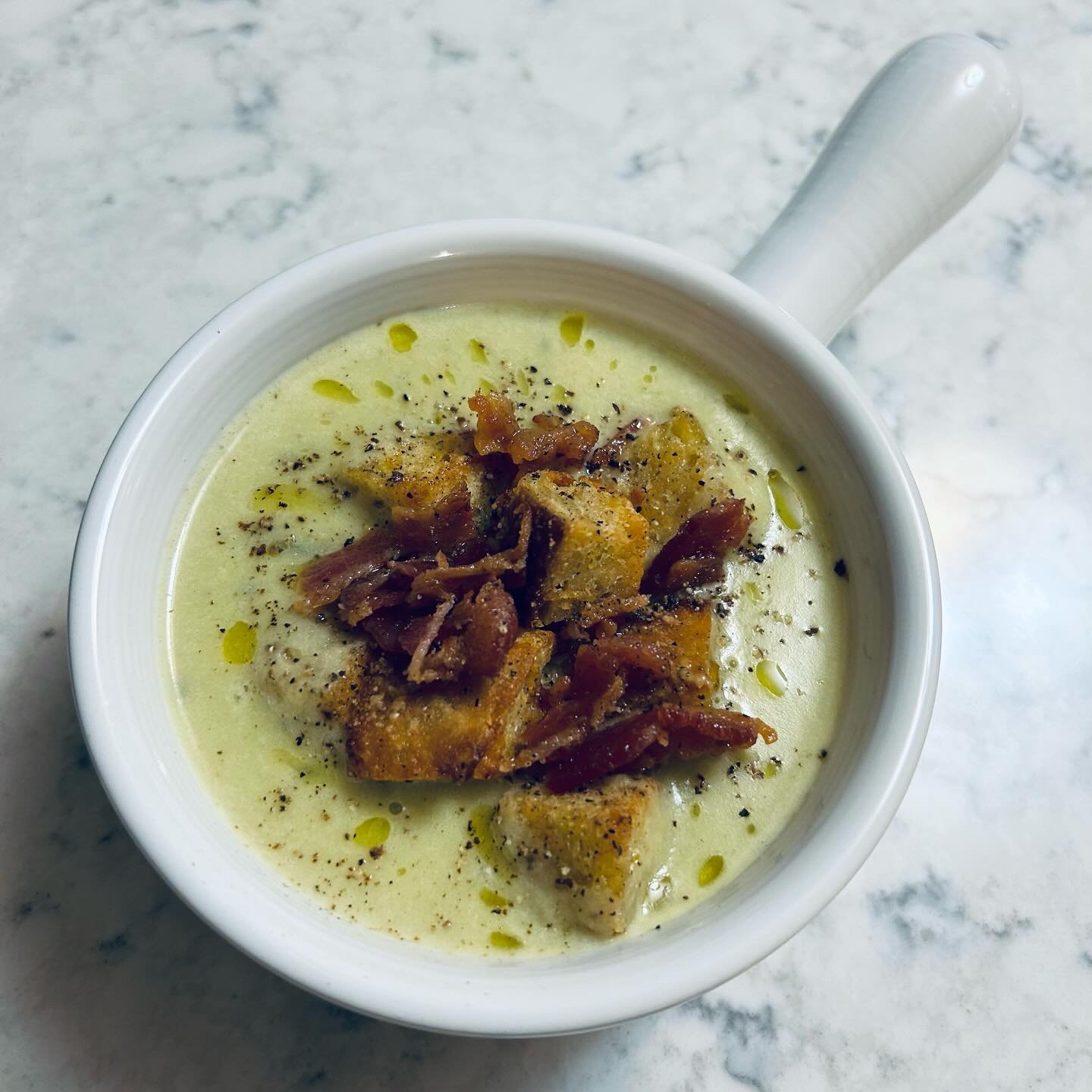 A mug of creamy soup topped with crispy bacon bits, black pepper, and drizzled with olive oil on a marble surface.