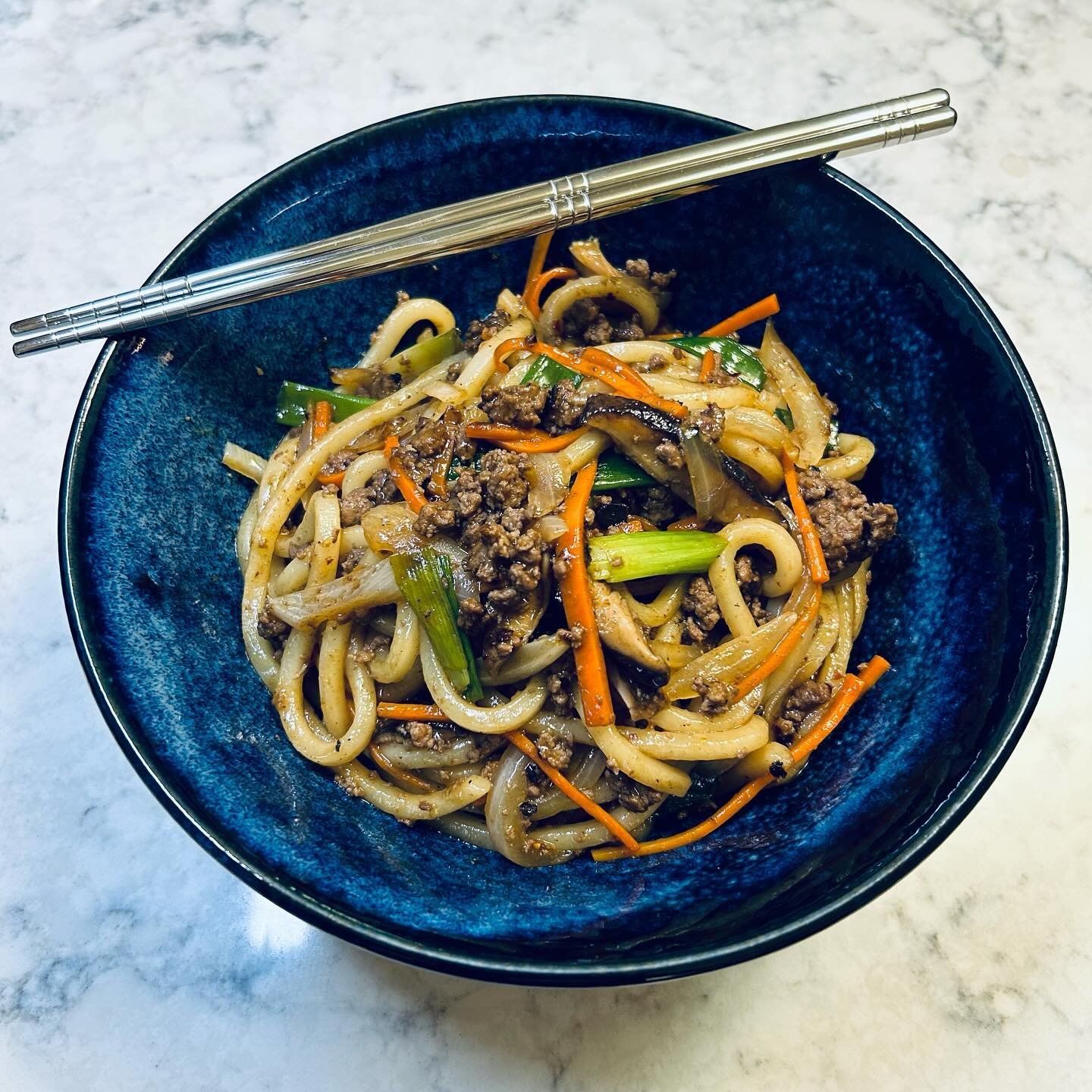 Bowl of stir-fried noodles with ground beef, carrots, green onions, and vegetables on a white marble surface, with chopsticks resting on the bowl's edge.
