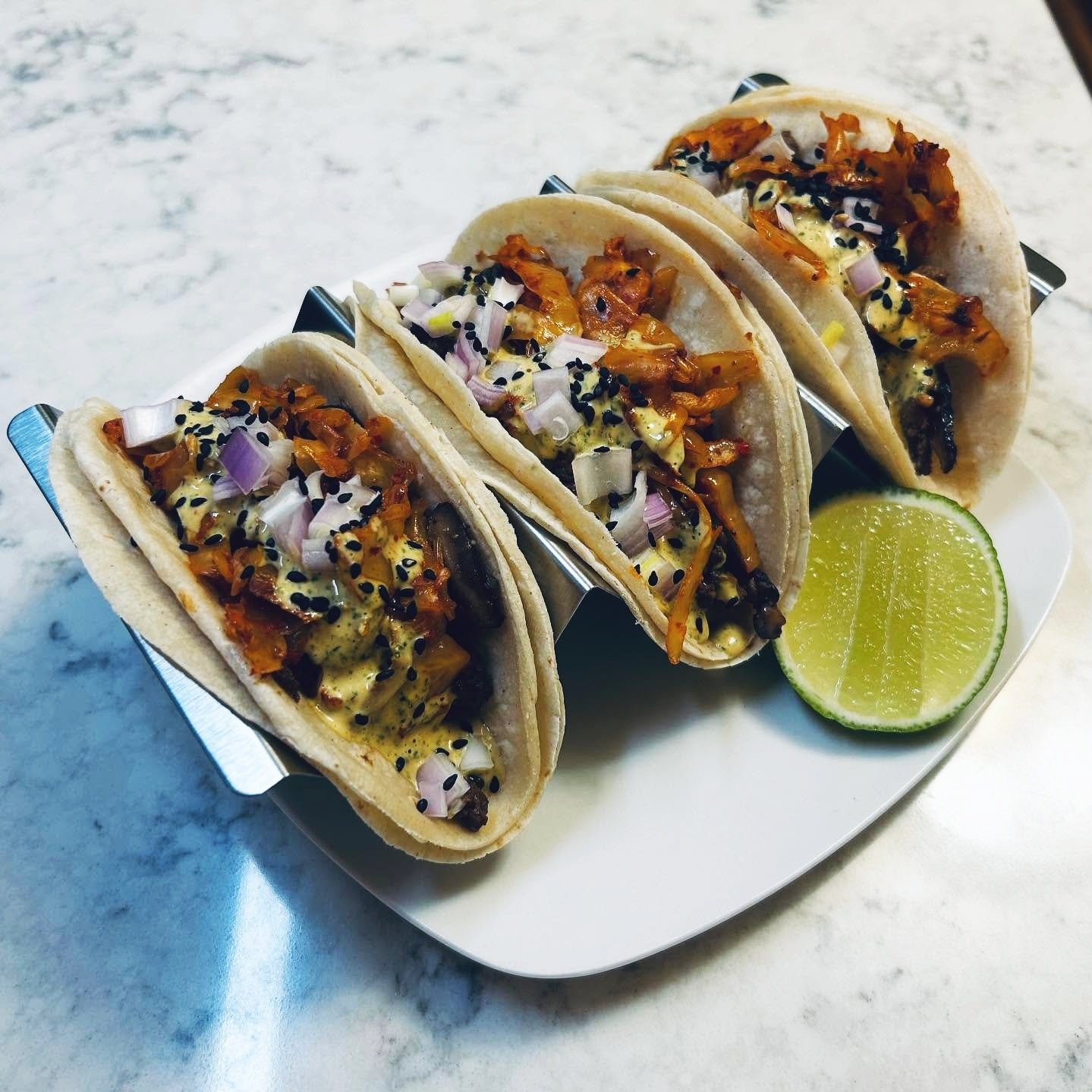 Four tacos filled with grilled meat, chopped onions, and sauce, garnished with black sesame seeds, served with a lime wedge on a white plate.