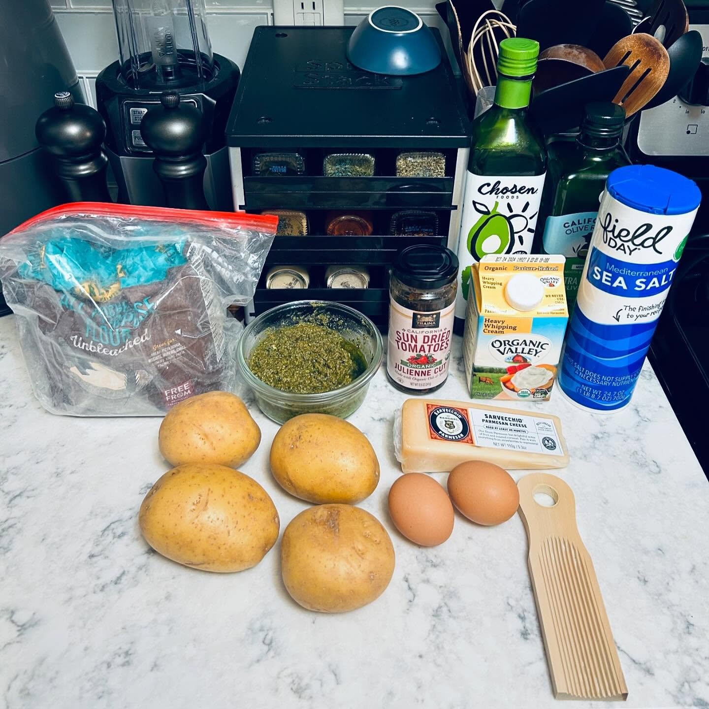 Ingredients for a recipe on a kitchen countertop, including potatoes, eggs, Parmesan cheese, sun-dried tomatoes, pesto, sea salt, heavy whipping cream, olive oil, a cutting board, and a grater.