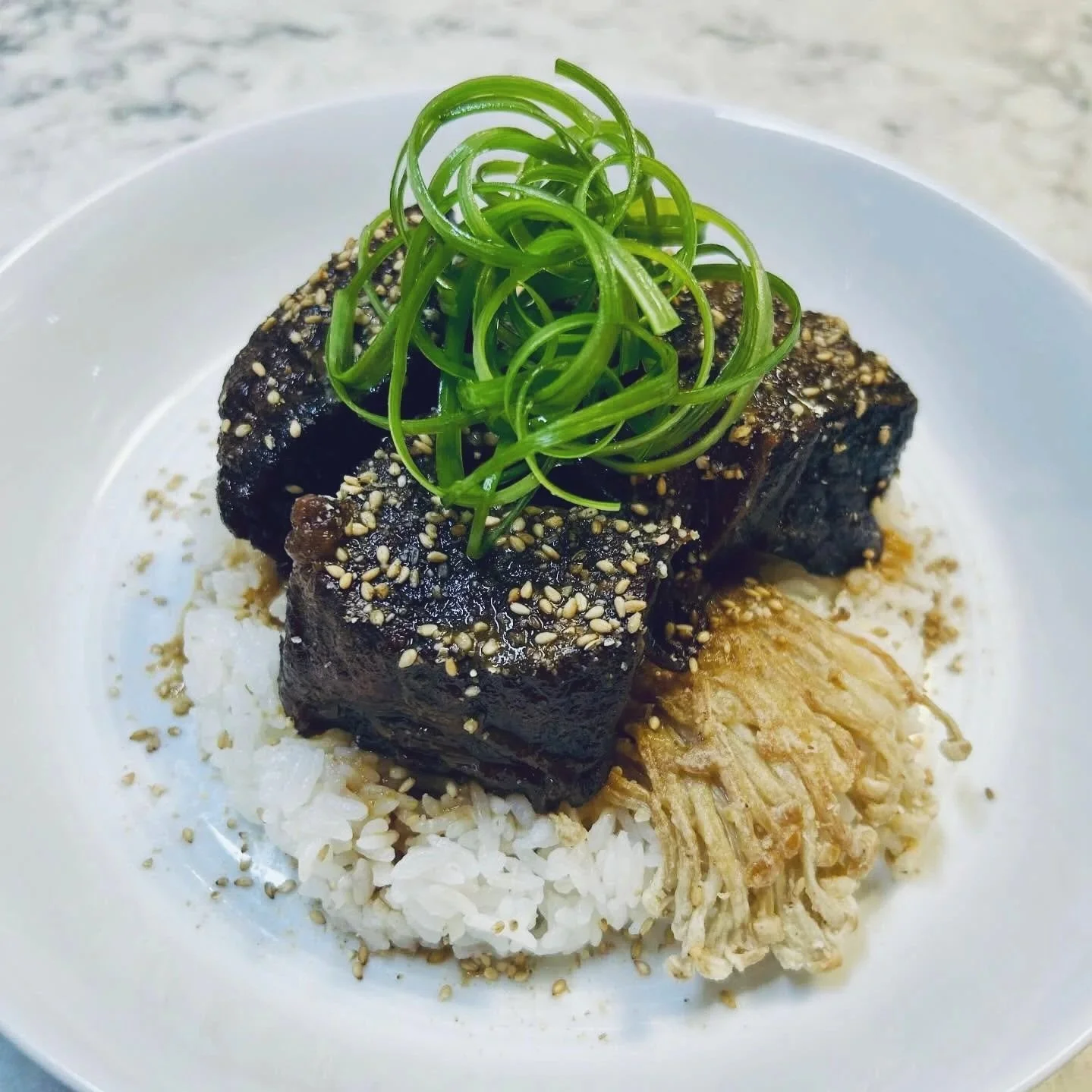 A white plate with rice topped with glazed beef, garnished with sesame seeds and green spiralized vegetable, and a side of shredded vegetables.