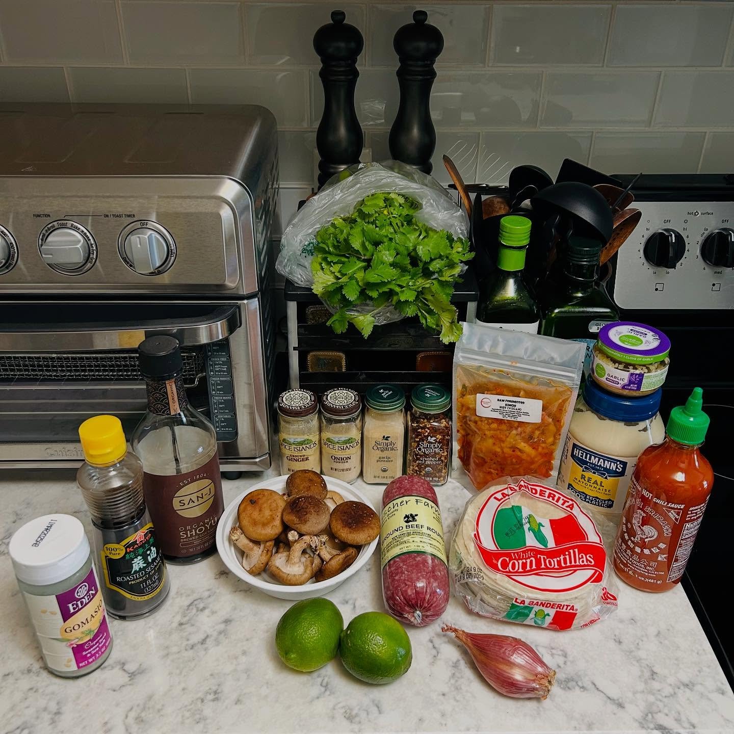 Countertop with fresh ingredients and cooking supplies including limes, mushrooms, cumin, onion, ground beef, kimchi, hot sauce, tortillas, and condiments, with an oven and kitchen utensils in the background.